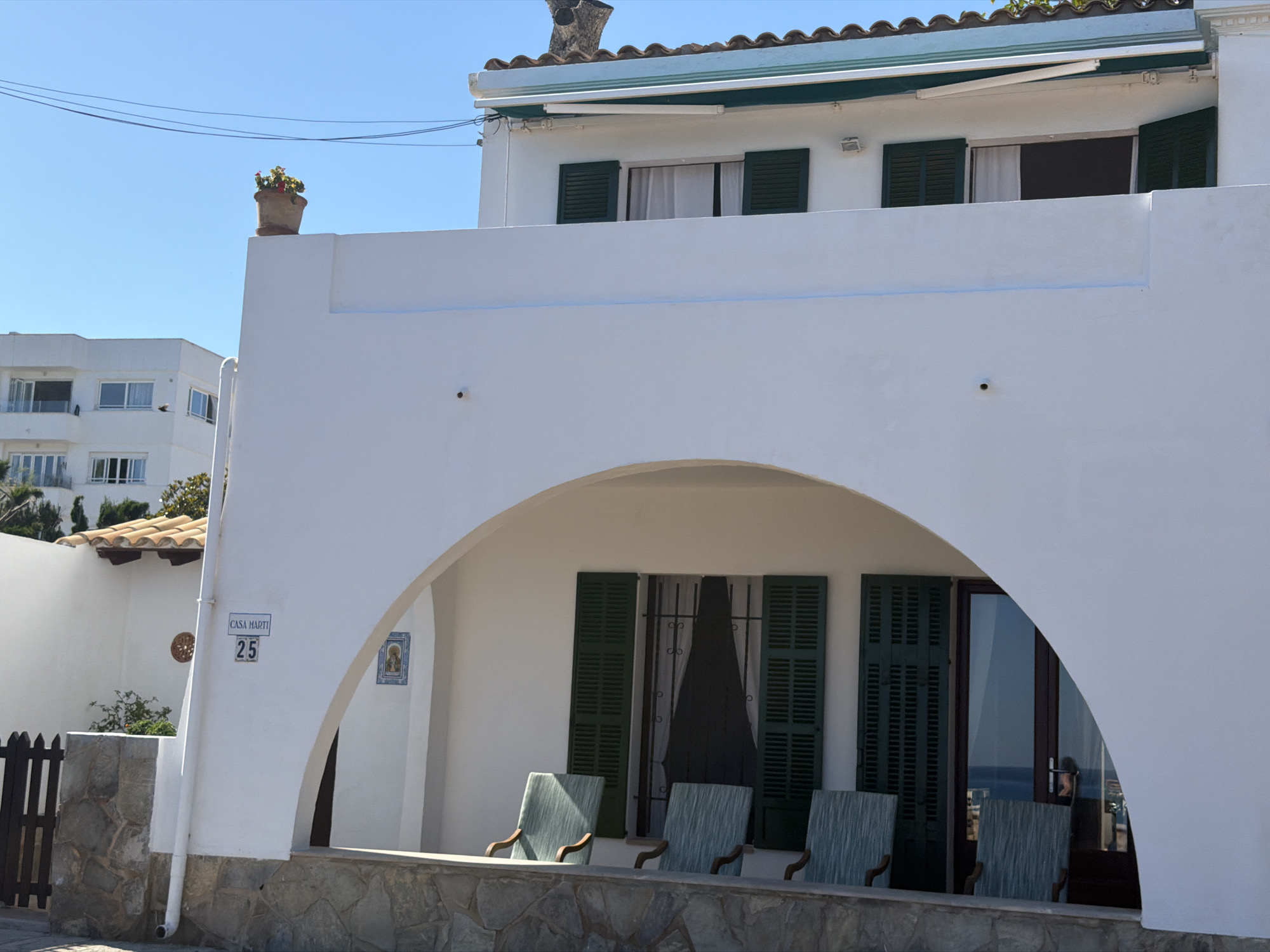 The image shows a white building in Capdepera, Spain, with a prominent arched entrance leading to a covered porch. On the porch are four chairs with wooden arms, positioned in front of two windows and a glass door, all featuring dark green shutters. The name "Casa Martí" and the number "25" are visible on a sign affixed to the building's exterior near the entrance. A flower pot sits atop the building's wall to the left. In the background, another white building is visible against a clear blue sky. Power lines run across the upper portion of the frame.