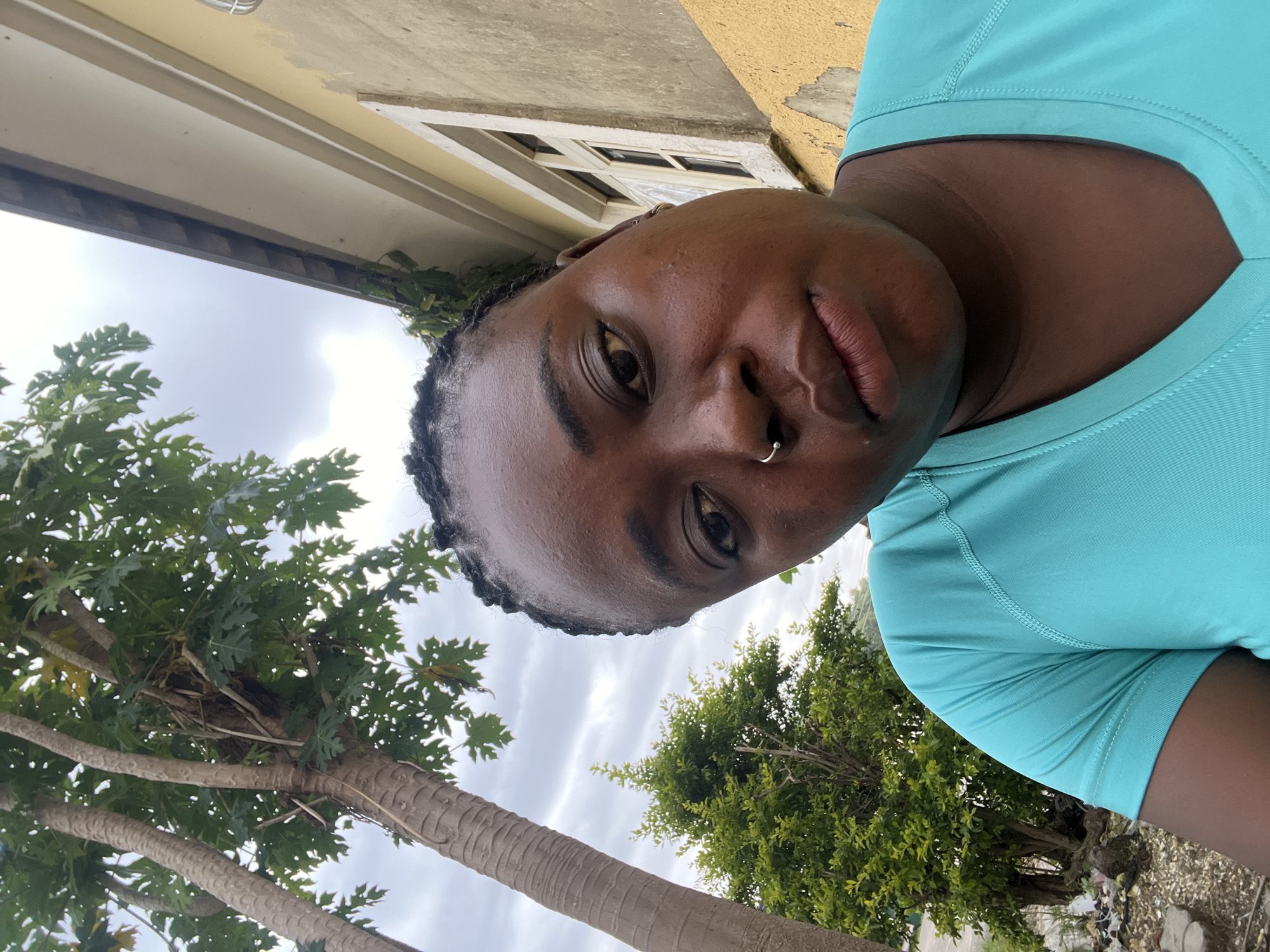 This is a close-up selfie of a Black woman wearing a turquoise t-shirt. She has her hair braided and is wearing a nose ring. She is outdoors with a tree and a building visible in the background. The building appears to be yellow with a white-trimmed window. The sky is overcast. The scene suggests a casual outdoor setting, potentially a residential area in Abuja, Nigeria.