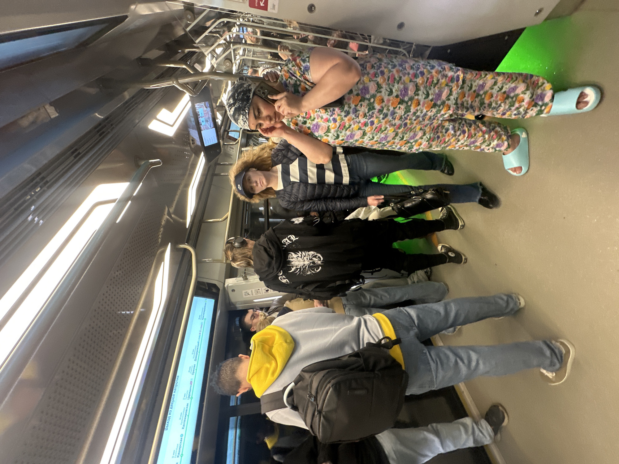 The image shows the interior of a public transport vehicle, likely a subway or train, in Moscow, Russia. Several people are visible, standing and sitting. In the foreground, a person wearing a light blue hooded sweatshirt with yellow accents and a dark backpack is seen from the back. To their left, another person in blue jeans is partially visible. Further up, a person with blonde hair wearing headphones and a black hoodie with a graphic print is standing.
In the upper part of the image, two women are more clearly depicted. One woman in a floral-patterned dress and light blue slides is on her phone. Next to her, a person with blonde, curly hair wearing a striped jacket and a blue baseball cap is looking towards the camera. The interior of the vehicle features overhead railings and illuminated screens displaying information. One screen clearly shows text in Russian, indicating "Exit from the doors opposite" and listing station names like "Kashirskaya," "Kantemirovoskaya," and "Domodedovskaya." The lighting suggests it is daytime. The overall atmosphere appears casual, with passengers engaged in their own activities.