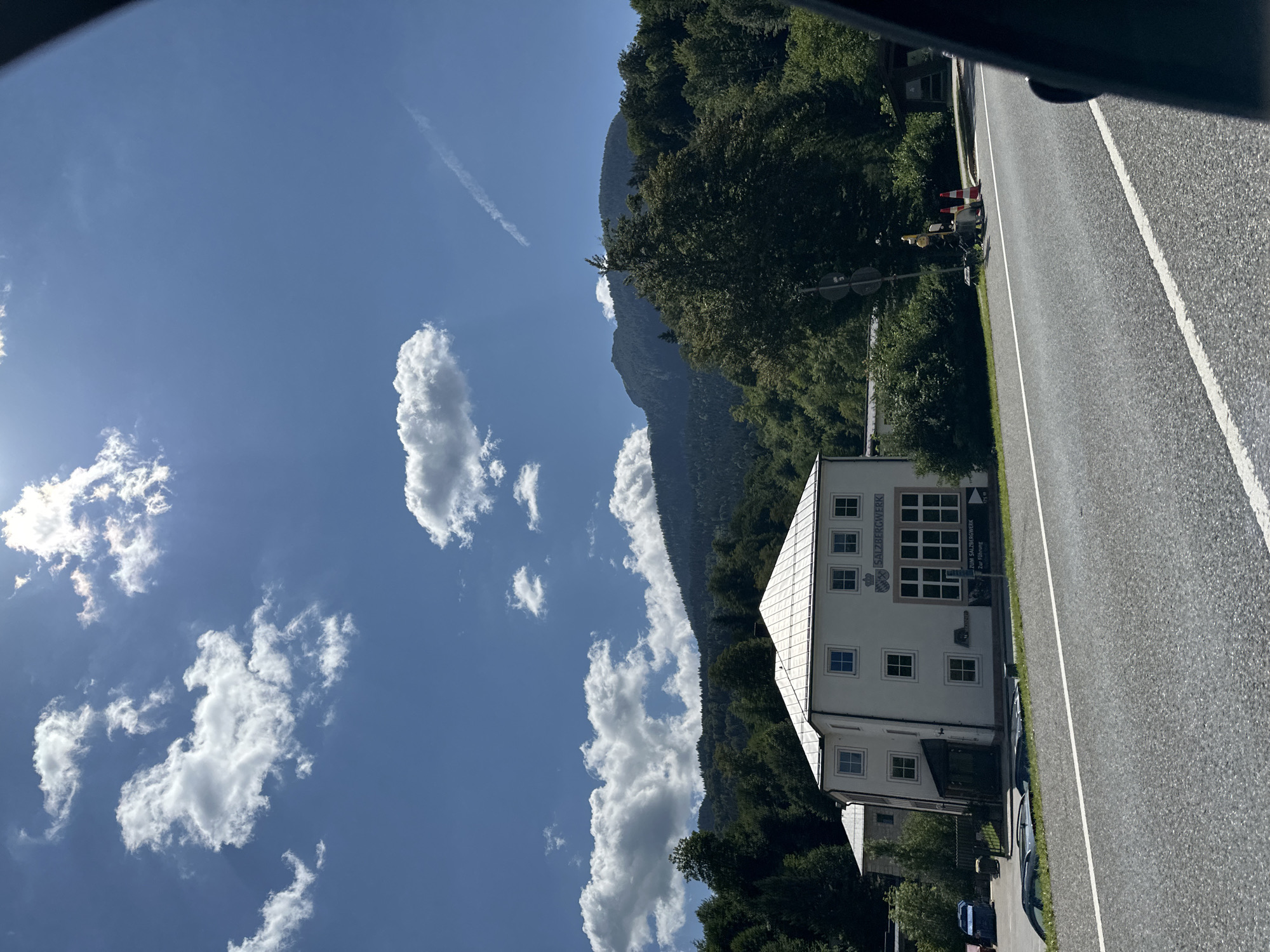 In Berchtesgaden, Germany, a two-story building with a white facade and a light-colored roof stands next to a road. The building has numerous windows and a sign that reads "SALZBERGWERK". Behind the building, a dense forest covers a mountainside, which transitions into a higher, more rugged mountain peak in the background. The sky is blue with scattered white clouds, and the sun is visible, casting light on the scene. The road in the foreground has white lane markings and appears to be a well-maintained asphalt surface. Traffic cones and a traffic signal are visible on the side of the road. Several cars are parked near the building.