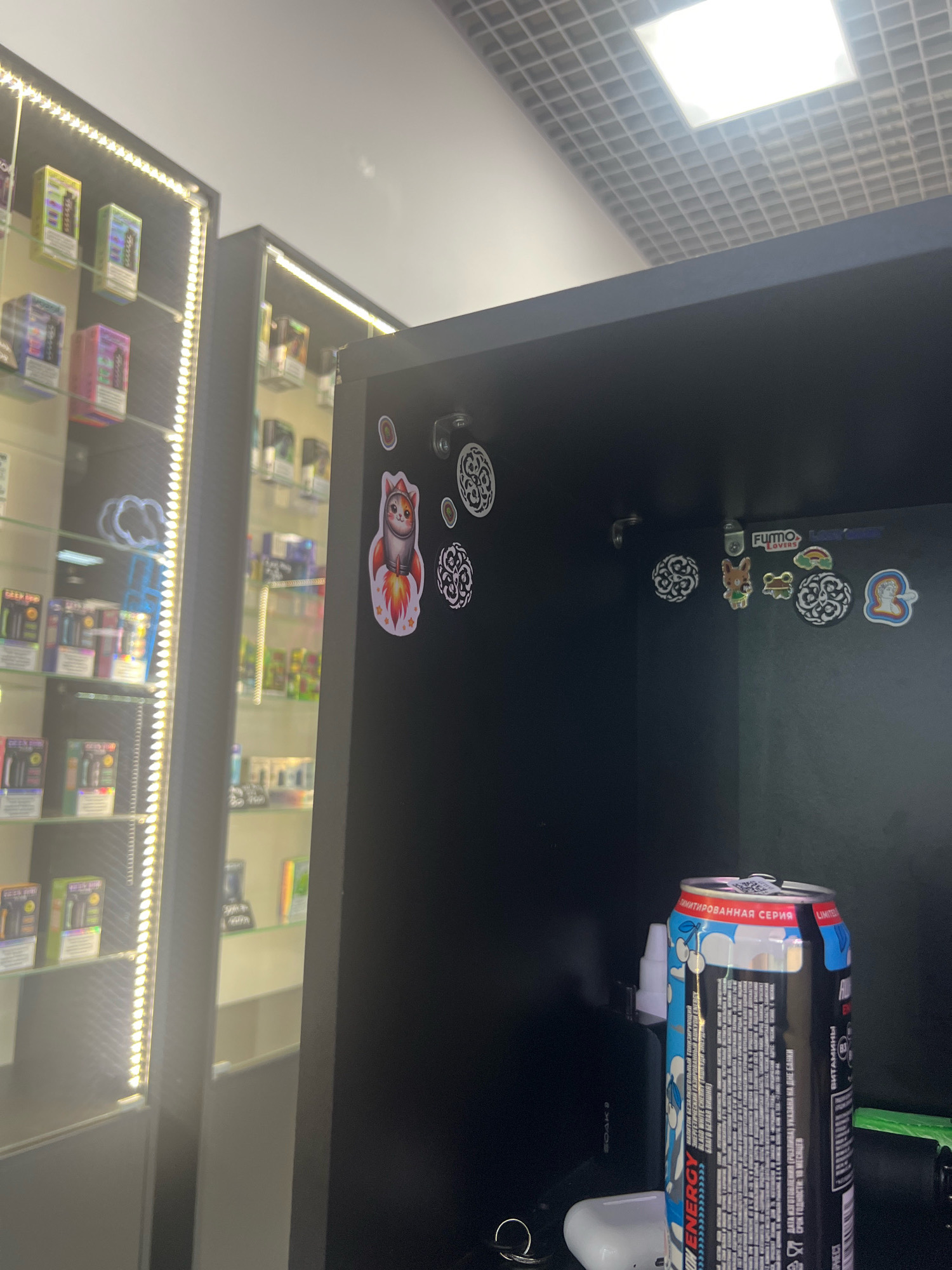 The image captures an indoor scene, likely a retail establishment specializing in electronic cigarettes and related products, based on the display cases filled with merchandise. The foreground is dominated by a black shelf or cabinet, upon which an energy drink can and white earbuds are placed. The energy drink can, with visible text in Russian, is positioned towards the right side of the frame. The background reveals a brightly lit display case filled with various packaged items, arranged on shelves. The overall lighting suggests an interior environment, possibly a shop, with artificial lighting illuminating the products. The ceiling features a grid of acoustic tiles with a bright light fixture. There is no indication of the specific location within Nizhny Novgorod, Russia, beyond the presence of Russian text on the energy drink.