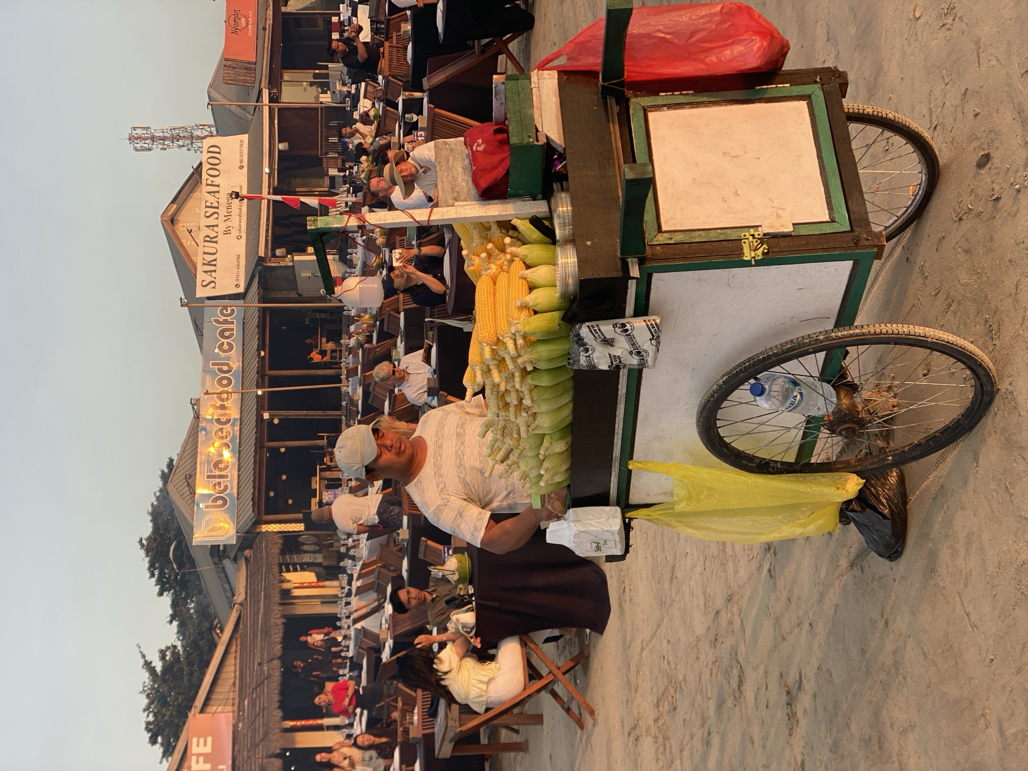 An outdoor scene in Kuta Selatan, Indonesia, captures a street vendor selling corn on a sandy beach in front of a row of seafood restaurants. The vendor, a man wearing a white t-shirt and a light-colored baseball cap, stands behind a cart laden with fresh corn cobs. The cart, which has two wheels and a wooden frame with green accents, also holds bags and other items. In the background, tables and chairs are set up on the sand, suggesting a dining area. Several people are seated at the tables, and others are walking around or standing. The restaurants have thatched roofs and signs with names like "Bela Seafood Cafe" and "Sakura Seafood." The sky is clear and the lighting indicates it is either early morning or late afternoon. The environment appears lively and suggests a tourist area where food vendors and restaurants cater to beachgoers.