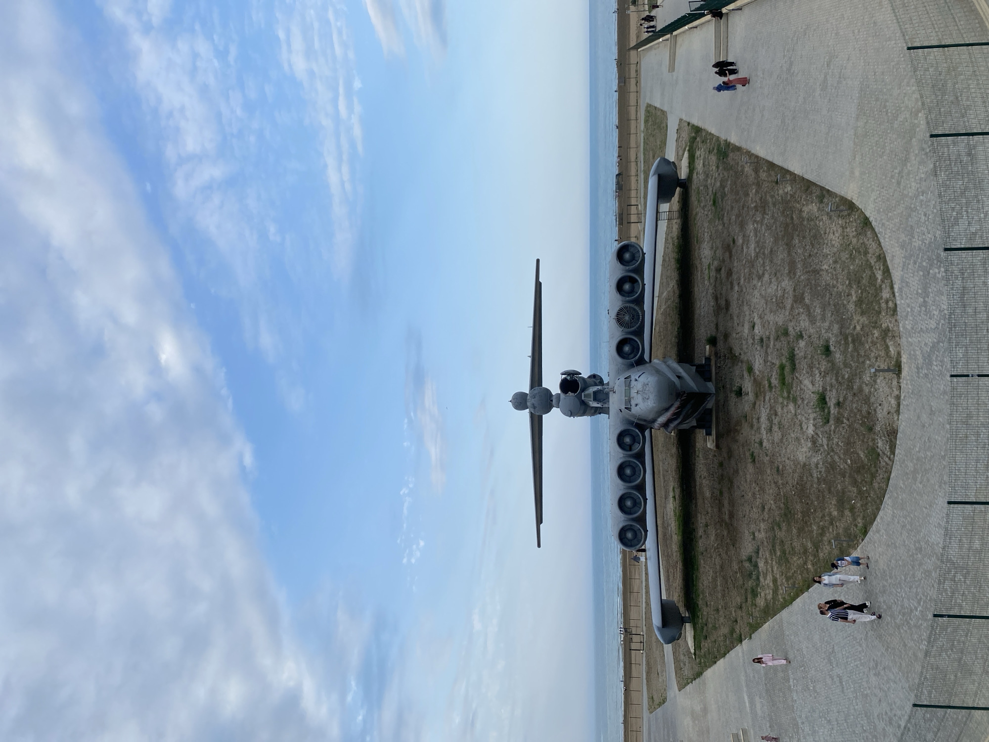 An aerial view captures a large, grey ekranoplan, an ekranoplan with eight engines mounted on its wings, displayed on a patch of dry grass near a paved walkway. The ekranoplan is positioned facing forward, with its large wings spread. Behind the ekranoplan, a calm, blue ocean stretches to the horizon, meeting a pale blue sky with scattered white clouds. Several people are walking on the paved pathway in the foreground, observing the ekranoplan. The environment appears to be a public space, possibly a park or museum, with a fence in the lower foreground. The location is identified as Arablyar, Russia.