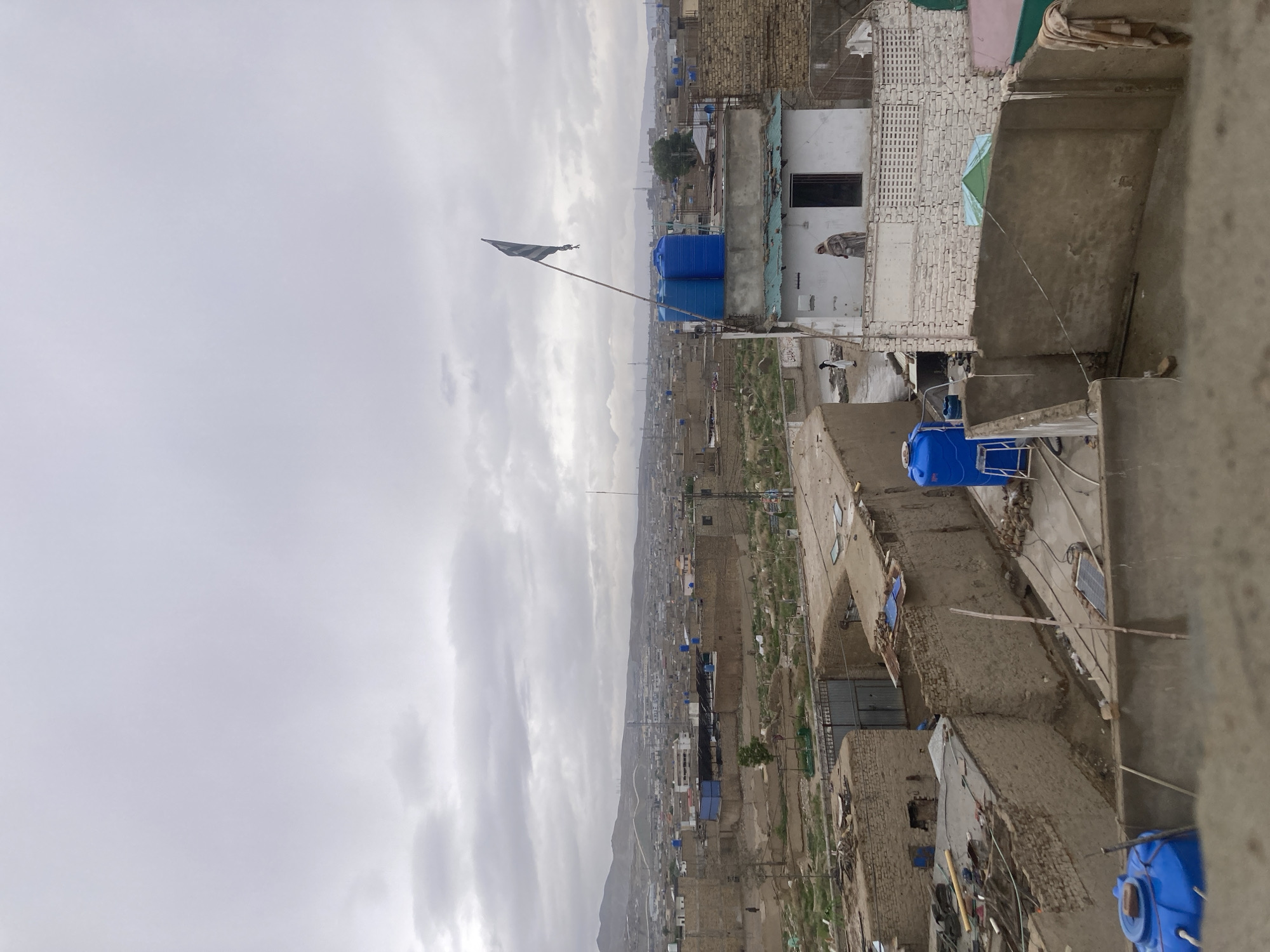 The image shows a cityscape of Quetta City Tehsil, Pakistan, viewed from a high vantage point, likely a rooftop. The foreground consists of buildings with rooftops cluttered with water tanks, solar panels, and various materials. In the midground, there are dense clusters of low-rise buildings extending into the distance, interspersed with open spaces and vegetation. A flag is visible on a tall pole amidst the buildings. The background features a mountain range under a cloudy sky. A figure is visible standing in a window in the middle ground. The scene depicts a densely populated urban area with a combination of residential and possibly commercial structures.