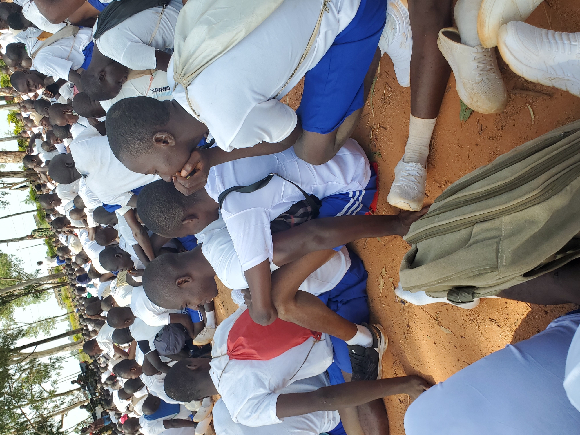 A high-angle, medium shot captures a gathering of young men, likely cadets, dressed in white t-shirts and blue shorts, seated on reddish-brown dirt ground under a canopy of sparse trees. The foreground is dominated by their legs and feet, clad in white socks and various athletic shoes. In the center, a cadet with a red drawstring bag slung over his shoulder is visible, his right arm resting on his thigh. To his right, another cadet, also in white and blue, is partially obscured, with his arms resting on his knees. The background reveals a larger group of similarly dressed individuals seated in rows, their faces turned in various directions, suggesting an organized assembly. A military vehicle is visible in the far distance, along with a few scattered buildings and more trees, hinting at an outdoor training or assembly ground, potentially within or near the city of Barakin Rafin Gora, Nigeria. The overall atmosphere appears calm and orderly, with the sunlight casting shadows on the ground.