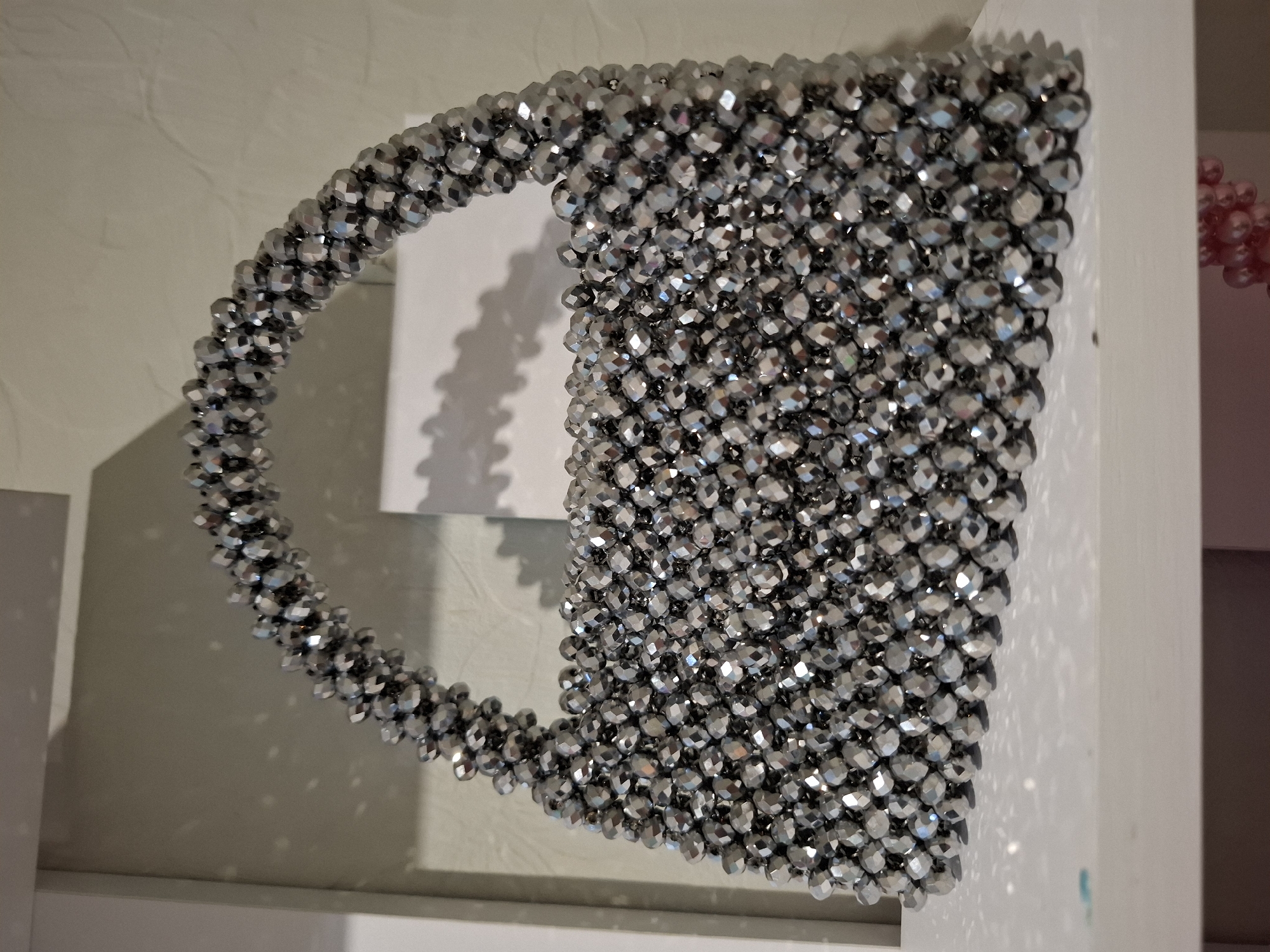 A silver beaded purse is placed on a white shelf. The purse has a curved handle and a rectangular body. The beads are faceted and reflect light, creating a shimmering effect. The background is a pale green wall with a shadow of the purse cast upon it. The shelf is clean and shows some minor imperfections. There is a hint of another object with pink beads visible below the white shelf. The scene appears to be indoors, possibly in a home or retail setting. There is no indication of the city of Korenovsk, Russia within the image itself.