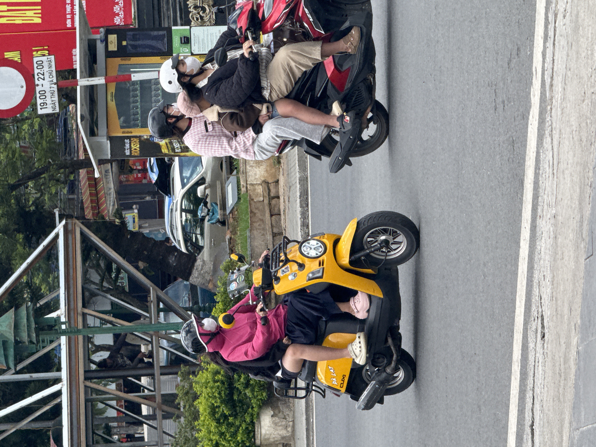 The image depicts a street scene in Da Lat, Vietnam, with two scooters in motion on a grey asphalt road.
On the left, a yellow scooter, bearing the text "DÀ LAT CLIXI," is ridden by two individuals. The driver, wearing a pink hooded jacket, black helmet, white face mask, and pink Crocs, steers the vehicle. A passenger in a black top, dark shorts, and beige Crocs sits behind the driver.
On the right, a red and black scooter carries three individuals. The driver, wearing a black helmet, white face mask, striped shirt, black quilted jacket, and light brown pants, operates the scooter. A child or small person in a pink cap, brown furry jacket, blue shorts, and beige Crocs sits in front of the driver. A rear passenger, wearing a black helmet, light pink and white checkered sweater, and light grey pants, sits behind the driver.
The background features commercial buildings and structures. To the far left, green foliage is visible. A multi-level metal framework, possibly part of a pedestrian walkway or building, stands with a person inside it. A tall green pole is situated near this structure. Further right, a white circular no-entry sign with a red border is present, below which a white rectangular sign reads "19:00 - 22:00 NGÀY THỨ 7 VÀ CHỦ NHẬT" (7 PM - 10 PM, Saturday and Sunday). A large red sign with white text, including "DAT LẠT," is visible on a building facade. A yellow "Rockstar" energy drink dispenser or advertising unit is also visible. Several cars are parked or in motion behind the scooters. The road surface has a white line marking its edge.
