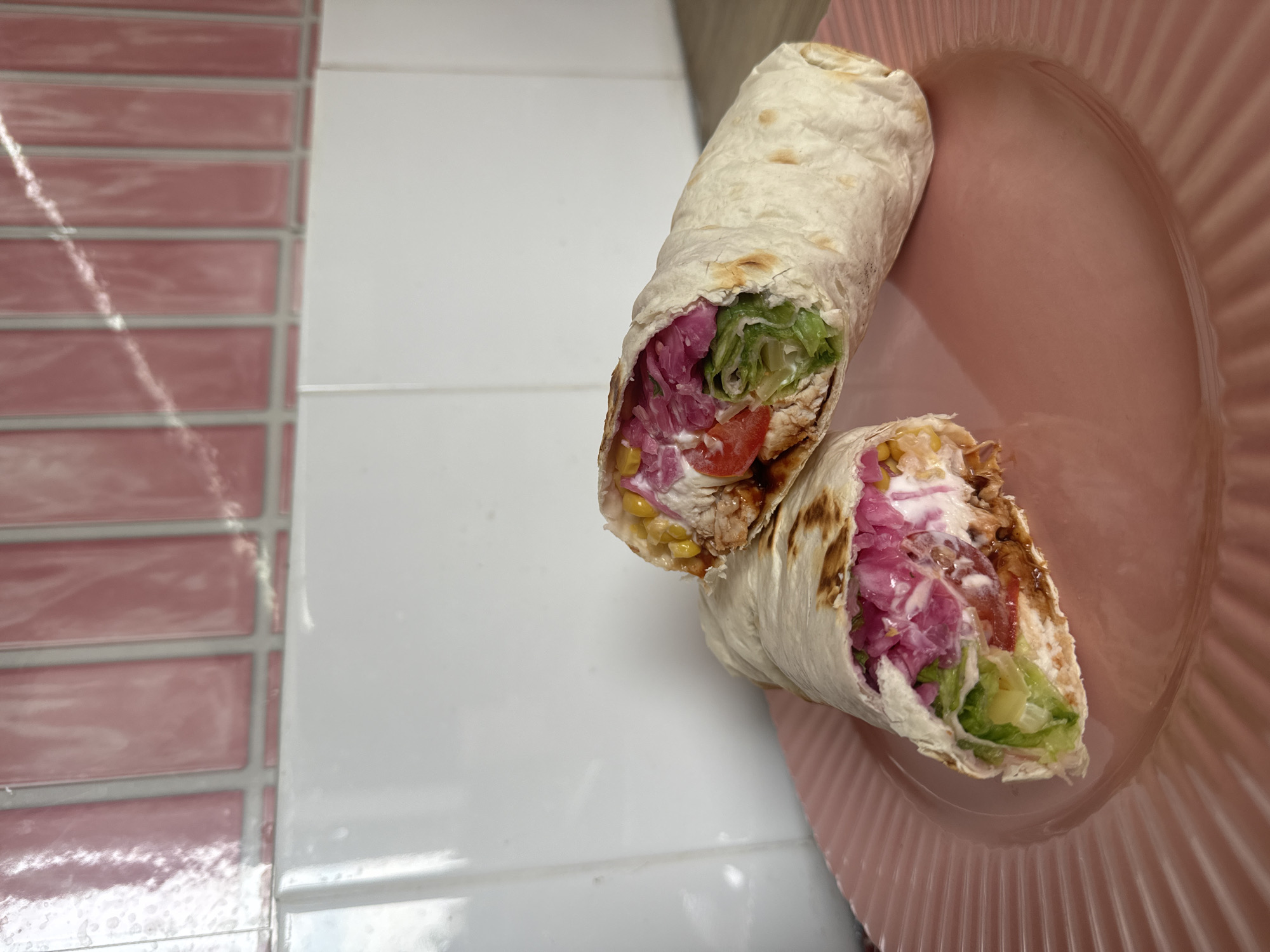 A close-up, eye-level shot captures two halves of a generously filled wrap resting on a pink, ribbed plate. The wrap, made from a lightly browned tortilla, is packed with ingredients that are visible through the open ends. These include shredded lettuce, sliced tomatoes, a creamy white sauce, vibrant purple pickled onions, and kernels of corn. The filling appears moist, with some dark sauce drizzled throughout. The background consists of a tiled wall, with the lower section tiled in white and the upper section in a row of elongated pink tiles. The image was taken in the city of Krasnoyarsk, Russia.