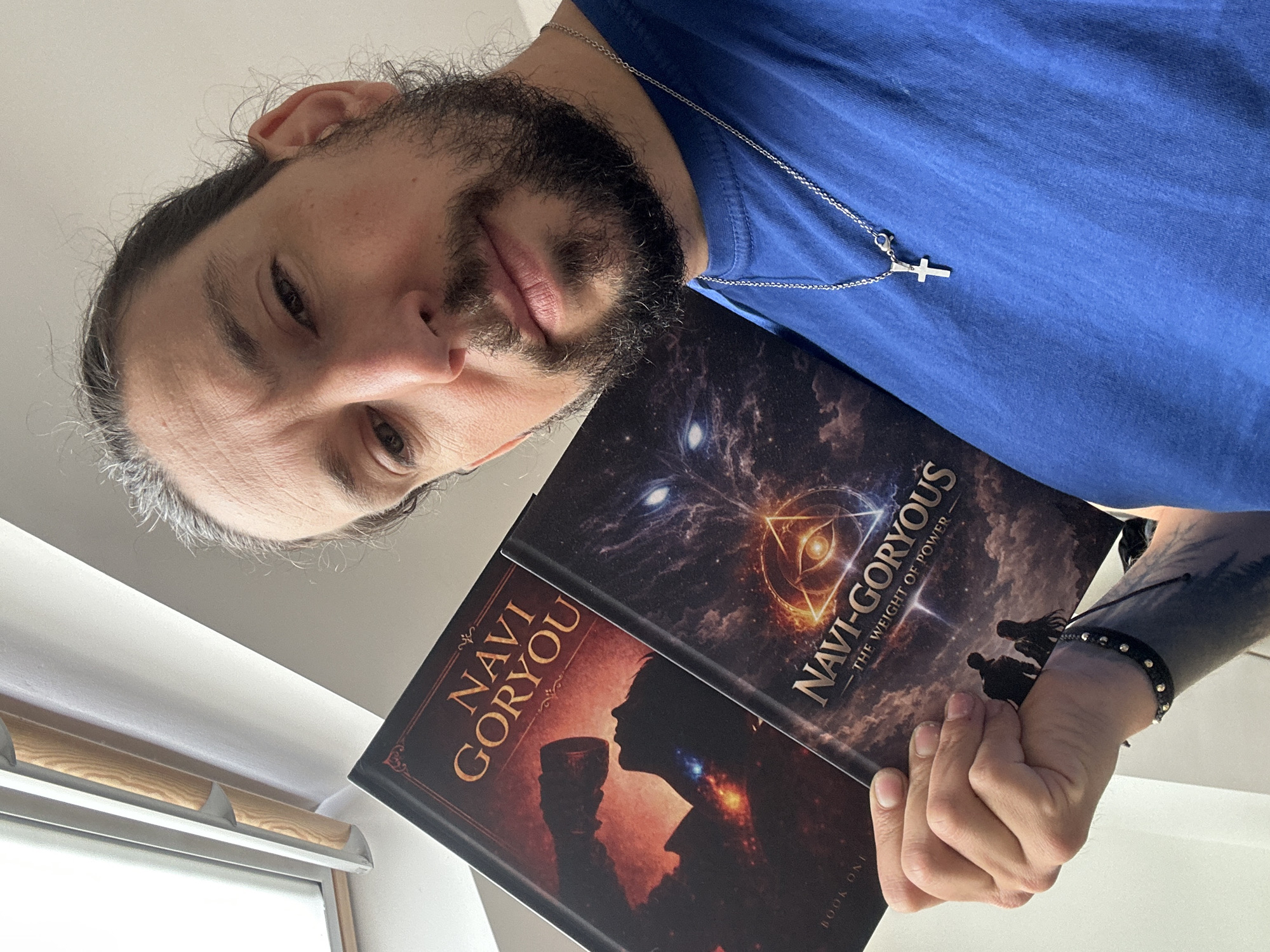 A man with a beard and long hair, wearing a blue t-shirt and a silver cross necklace, is holding two books titled "Navi-Goryou" in his hands. The book covers are visible, displaying artwork. The man has a tattoo on his arm and is wearing a beaded bracelet. The photo appears to be taken indoors, possibly near a window, as light is visible from the left side. There is no indication of the location being Nivelles, Belgium in the image.