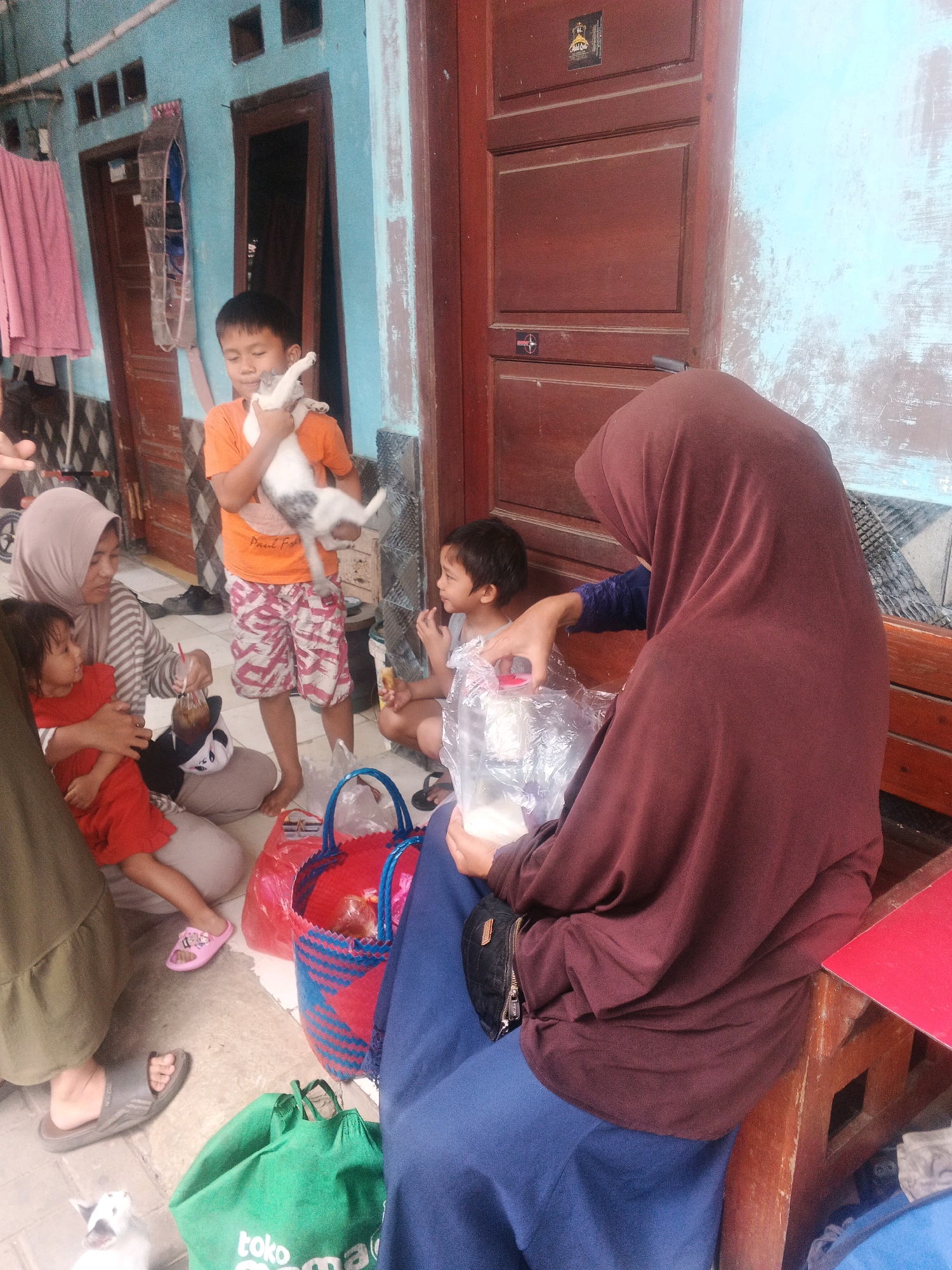 The image depicts an outdoor scene in the city of Kadu, Indonesia, in front of a building with a light blue painted exterior and a dark brown wooden door. In the foreground, a young boy in an orange t-shirt and patterned shorts holds a white and grey cat. To his left, a woman wearing a hijab and a striped shirt kneels, holding a young child in a red dress. Next to them, a woman in a dark red hijab and blue pants is seated on a wooden bench, holding a plastic bag with contents. Several bags are on the ground, including a red and blue woven basket and a green reusable bag. A small white cat is visible on the ground to the lower left. The scene appears to be a casual gathering, possibly in front of a residential dwelling.