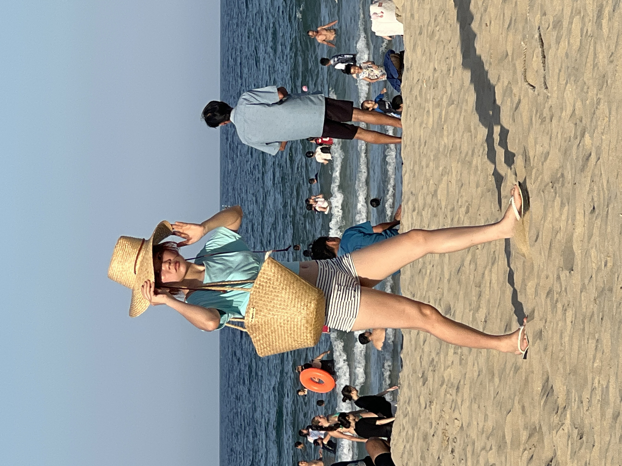 An eye-level medium shot captures a young woman in the foreground walking on a sandy beach. She is wearing a light blue short-sleeved shirt and black and white striped shorts. On her head, she sports a wide-brimmed straw hat, which she is adjusting with her hands. A woven straw bag hangs from her right shoulder. She is walking to the left, casting a shadow on the sand.
Behind her, the sea is filled with numerous people swimming and wading in the water. The water is a deep blue, with gentle waves breaking near the shore. In the middle ground, a man in a light blue shirt and dark shorts stands facing away from the camera, looking out at the sea. Several other beachgoers are visible in the water and on the sand, engaged in various activities. The overall atmosphere suggests a sunny day at a popular beach location.
This scene is consistent with the vibrant beach life found in Đà Nẵng, Vietnam, a city renowned for its long stretches of sandy coastline and bustling seaside activities.