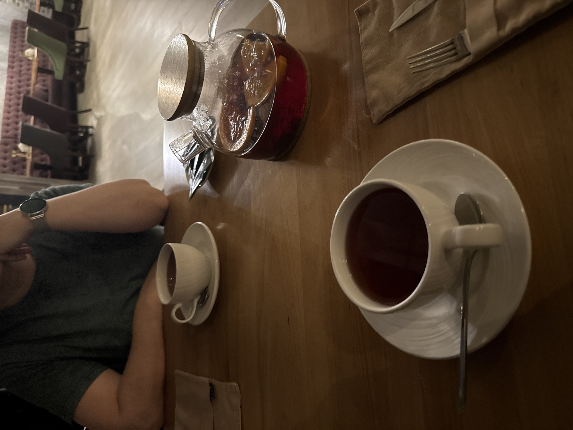 The image shows a person seated at a wooden table in what appears to be a restaurant or cafe. The person is wearing a dark green shirt and a watch. On the table, there is a glass teapot filled with a red beverage containing fruit slices, two white teacups with saucers and spoons, and a tan napkin with cutlery. The location is unspecified, and there is no verifiable connection to Uryupinsk, Russia. The focus is on the beverages and table setting.