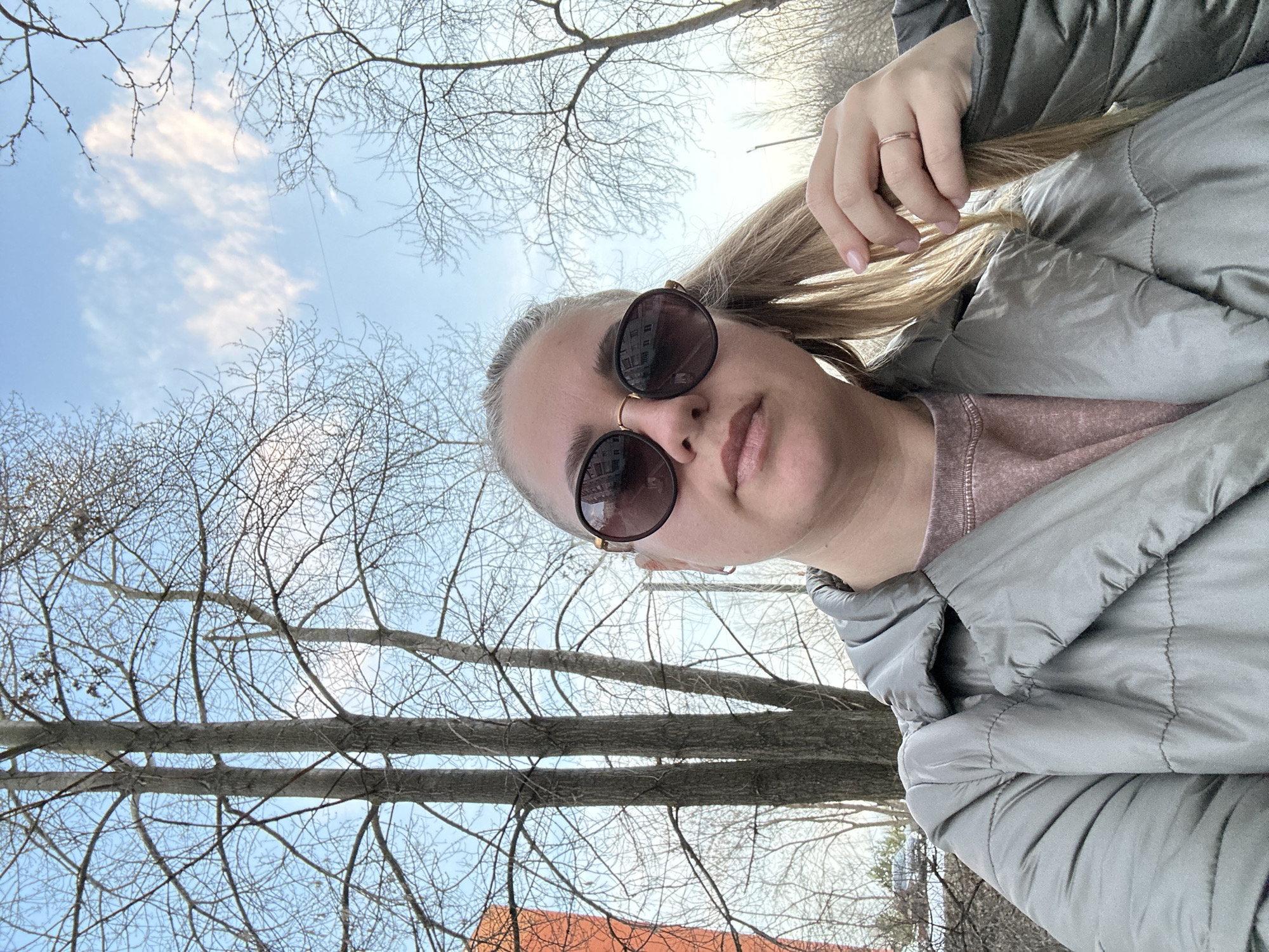 A selfie-style, low-angle photo shows a woman with blonde hair and sunglasses, wearing a gray puffy jacket. The background features bare trees against a blue and slightly cloudy sky. A glimpse of a reddish-orange roof is visible in the lower left. The location is Chaikovsky, Russia. The woman is holding her hair with her right hand. She is wearing a ring on her right hand. She is also wearing a nose ring. The sunglasses reflect a building.