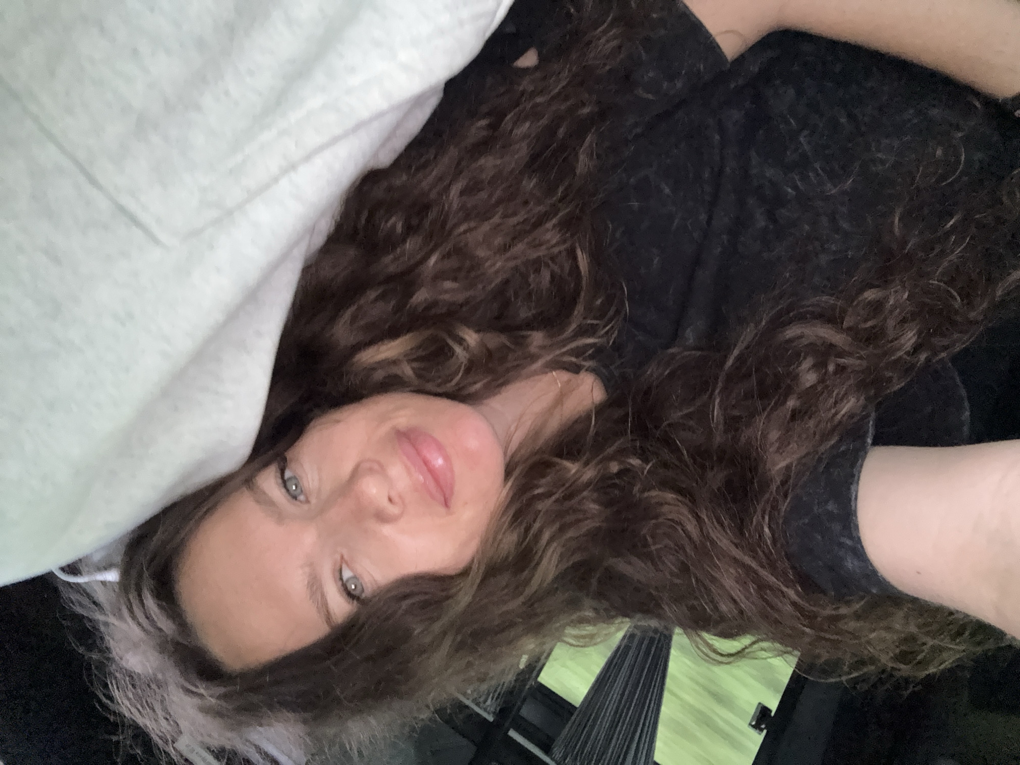 A medium close-up selfie shows a woman with long, wavy brown hair, light blue eyes, and light skin. She is looking at the camera and smiling slightly. She is wearing a dark t-shirt. A light-colored blanket or sheet is draped over her head and shoulder. The background appears to be inside a vehicle, with dark tinted windows showing a blurred green outdoor scene. The image quality is slightly grainy. The selfie was taken in the city of Turbaza Katun', Russia.