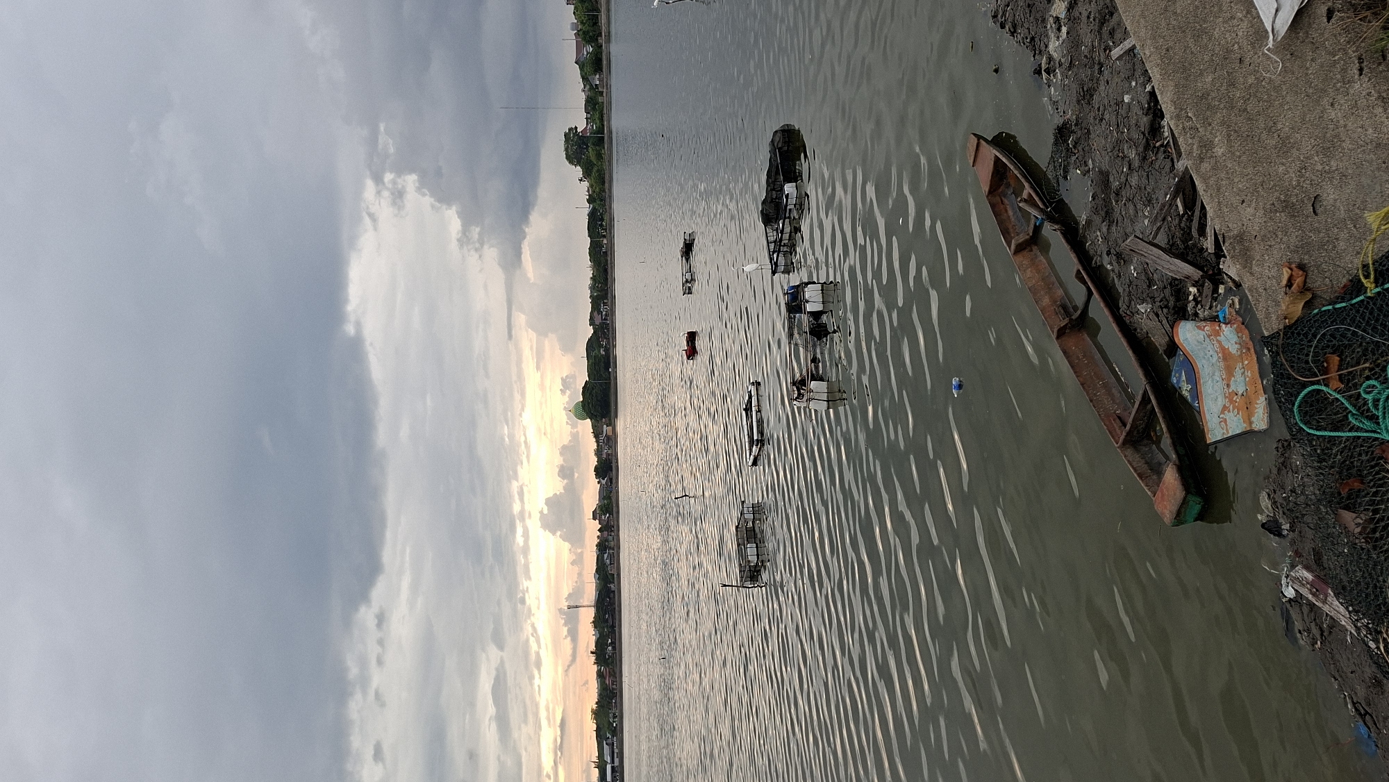 The image depicts a waterfront scene in Lhokseumawe, Indonesia, likely near the mangrove forest area. The sky is overcast with a mix of gray and lighter clouds, suggesting a late afternoon or early morning setting, with a hint of sunset colors on the horizon. The water is calm, with small ripples on its surface.
In the water, several floating structures, possibly fish farms or cages, are visible. Two white birds, likely egrets, are perched on these structures. A lone, small, rusty wooden boat is partially submerged at the edge of the water, resting on the muddy shore. Debris, including plastic waste, fishing nets, and discarded materials, is scattered along the embankment.
In the background, across the water, a shoreline with buildings, trees, and a prominent green domed structure, possibly a mosque, can be seen. A telecommunications tower is also visible. The overall impression is one of a utilitarian waterfront, potentially used for aquaculture and fishing, with signs of pollution and neglect.