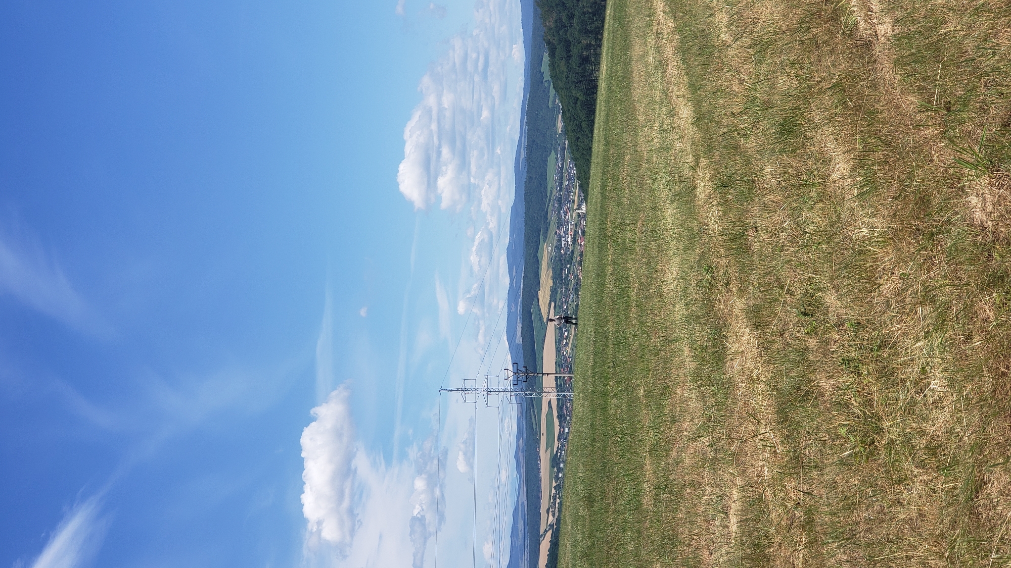 The image depicts a scenic view from a grassy hillside overlooking a town nestled in a valley. The sky is a vibrant blue with scattered white clouds, some appearing wispy and others more cumulus-like. In the foreground, a dry, mown field of grass extends across the frame.
Dominating the mid-ground to the left is a tall, metallic electricity pylon with several wires extending horizontally. A solitary figure, appearing to be a person wearing a light-colored top and darker pants, stands on the hillside to the right of the pylon, facing away from the camera and looking towards the town.
The town itself, located in the valley, features numerous buildings with red-tiled roofs, interspersed with patches of green trees. A white church steeple is visible within the town. Beyond the town, rolling hills covered in a mix of green and yellow vegetation rise, transitioning into darker, more densely forested hills in the distance, forming a layered mountainous backdrop against the horizon. The overall impression is one of a peaceful, rural landscape on a sunny day. This scene is consistent with views from the hills surrounding Prešov, Slovakia, which are known for their scenic vistas of the city and surrounding countryside.
