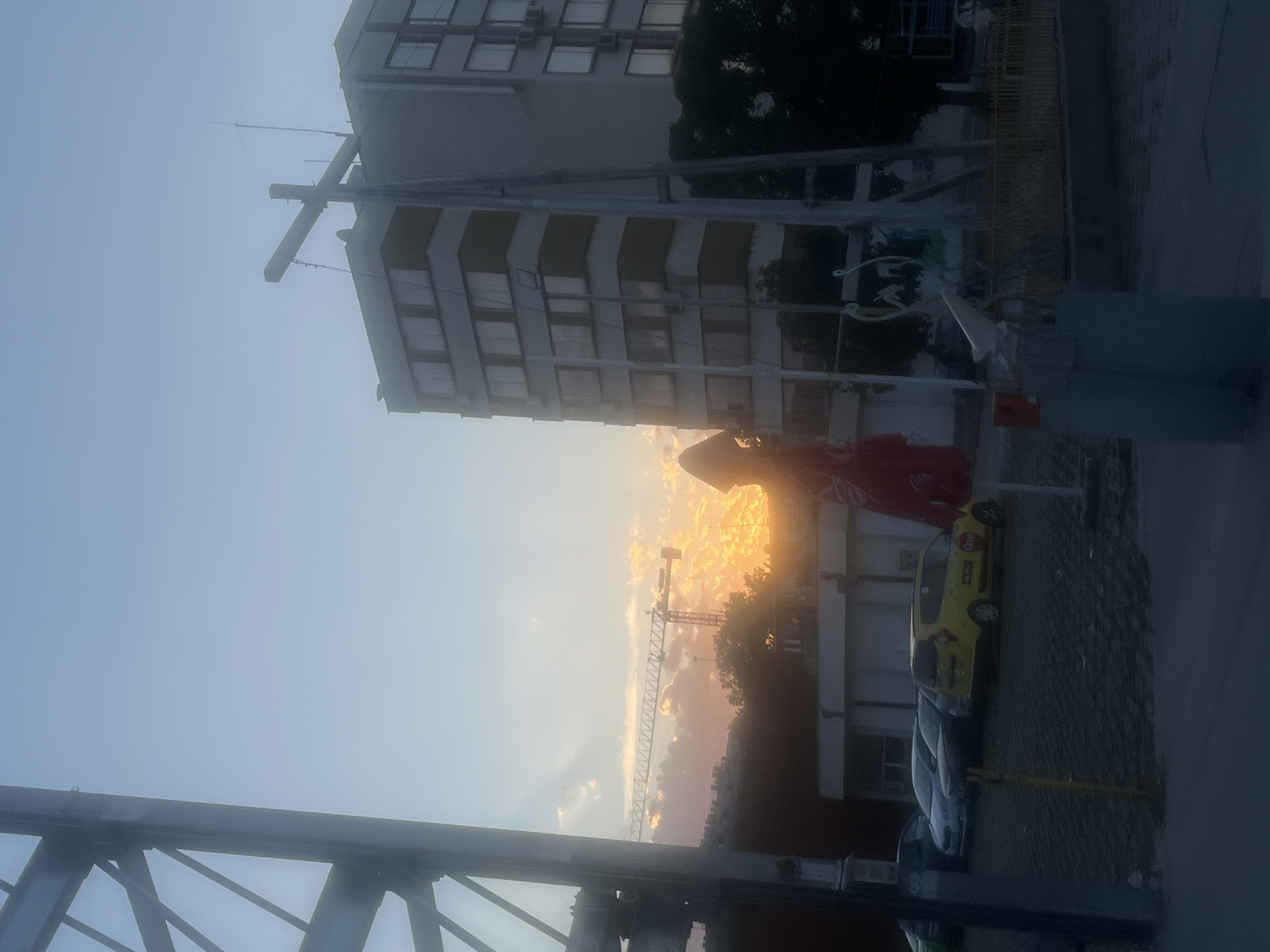The image depicts a street scene with buildings, vehicles, and a construction crane against a sky with a sunset. In the foreground, a large metal structure is visible on the left, obscuring part of the view. To the right, a tall, multi-story building stands, with its facade showing a pattern of windows and what appear to be air conditioning units. In front of this building, a street with parked cars and a yellow taxi is visible. A construction crane is seen in the midground, silhouetted against the vibrant orange and yellow hues of the sunset. The sky is a gradient of blue and warm sunset colors, with some scattered clouds. There are no visible people in the image. The overall atmosphere is somewhat hazy, likely due to the sunset and possibly atmospheric conditions.