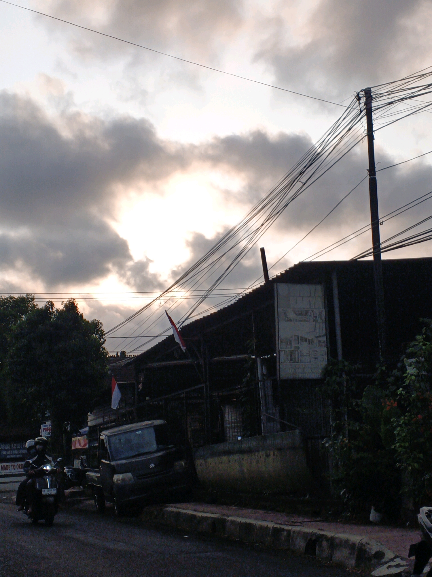 In Sukawati, Indonesia, on a cloudy day with the sun breaking through the clouds, a dark, utilitarian pickup truck is parked on the side of a paved road, its front wheels partially on the curb. To the left of the truck, a motorcycle with two occupants is approaching, the driver wearing a helmet and the passenger behind. The motorcycle has its headlights on. Overhead, a complex network of electrical wires crisscrosses the sky, supported by wooden poles. Buildings with dark corrugated roofs line the street, with one building displaying a faded advertisement. An Indonesian flag, red and white, is visible on a flagpole near the building. Green foliage and trees are present, some appearing overgrown, adding to the street's character.