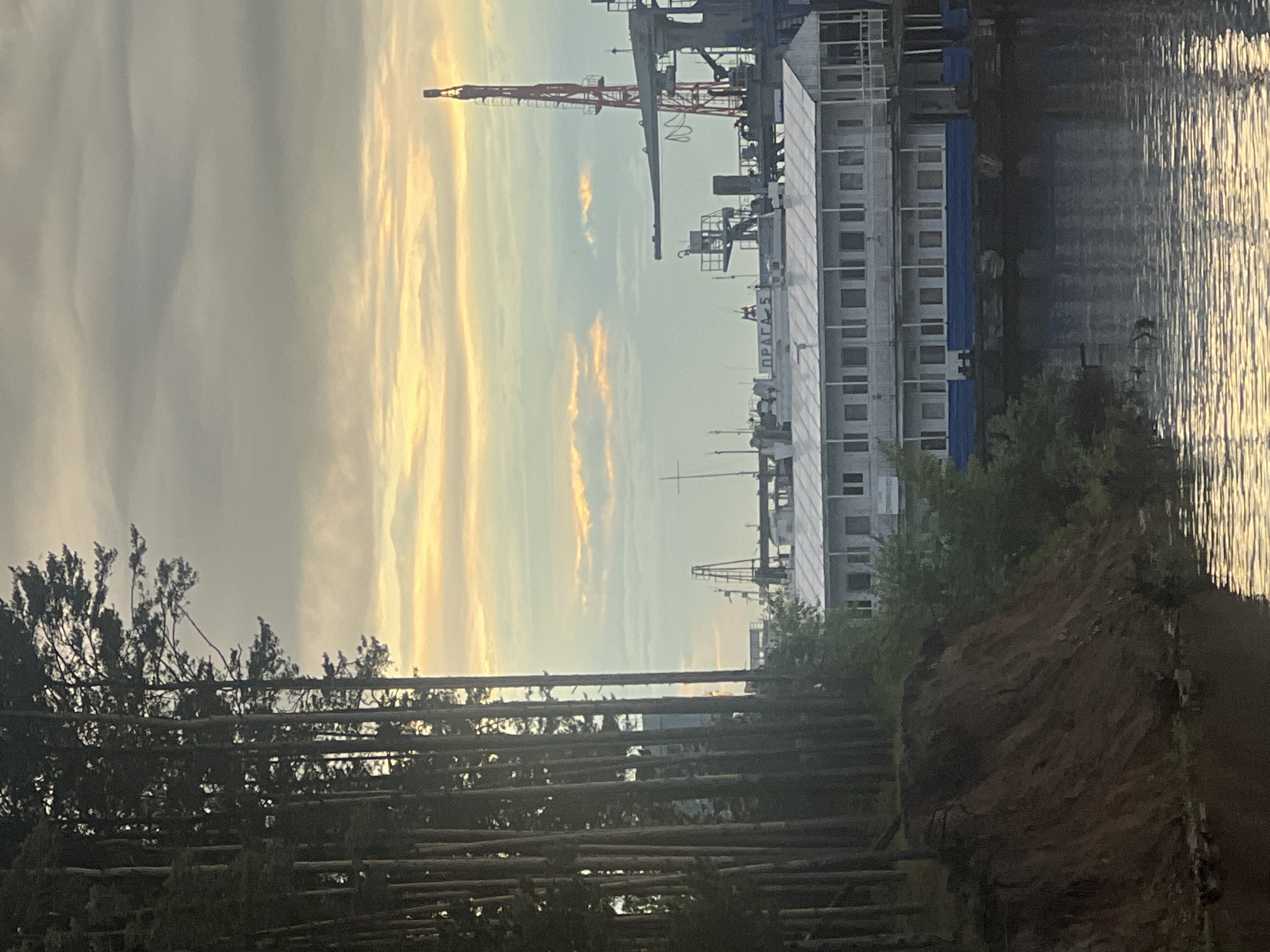 This media file depicts a tranquil scene at what appears to be a port or industrial riverside in Chaikovsky, Russia, likely during sunrise or sunset. The image is composed of distinct horizontal layers: a tree-lined bank on the left, a body of water in the middle, and industrial structures with a golden sky on the right.
In the foreground on the left, numerous felled trees or logs are stacked horizontally, forming a textured barrier. Behind these, a dense cluster of tall, dark-foliaged trees stands, obscuring the horizon. A patch of dark brown soil or earth is visible on the bank leading down to the water.
The central part of the image features a wide body of water, possibly a river or lake. Its surface shimmers with golden reflections, mirroring the vibrant light from the sky, suggesting calm conditions.
On the opposite bank, a large, multi-story white structure, resembling a ship or a dockside building, dominates the right side of the frame. Several industrial cranes are visible behind and above this structure, including a prominent red crane. On the side of the white structure, the Cyrillic text "ПРАГА-5" (PRAGA-5) is clearly visible. This could denote the name of a vessel or a building.
The sky, occupying the upper portion of the image, is adorned with elongated clouds streaked with warm golden, yellow, and soft blue hues, indicating the soft, ambient light of either early morning or late evening. No people are visible, and the scene is static, evoking a sense of quiet solitude. The overall impression is one of an industrial landscape at a peaceful time of day.