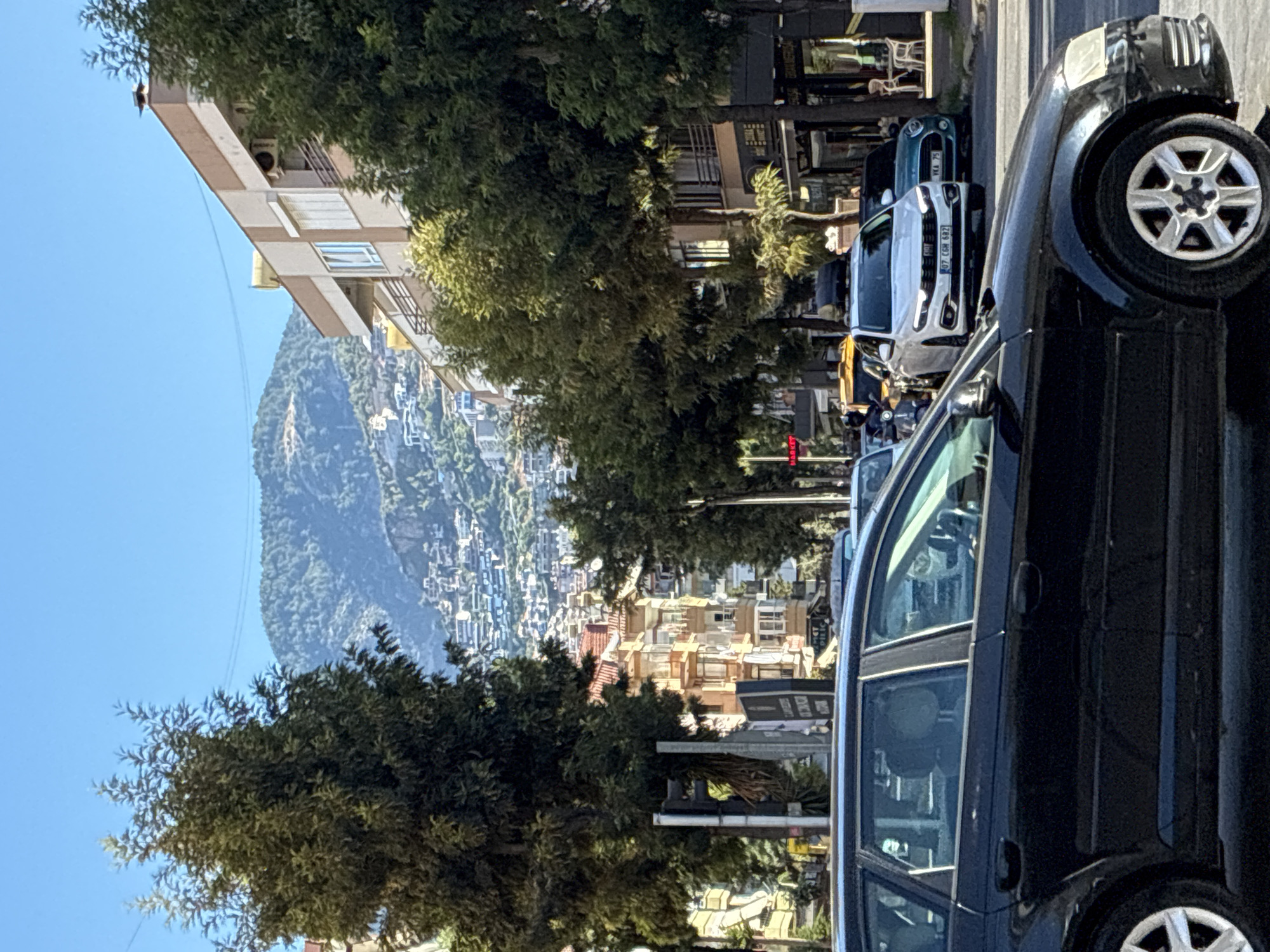 This is a street scene in Alanya, Turkey, featuring a view of a mountain covered in buildings in the background. In the foreground, a black car is parked on the right side of the road. The street is lined with trees and multi-story buildings. Several other vehicles, including a white Fiat and a teal MINI Cooper, are visible on the road. The sky is clear and blue.
