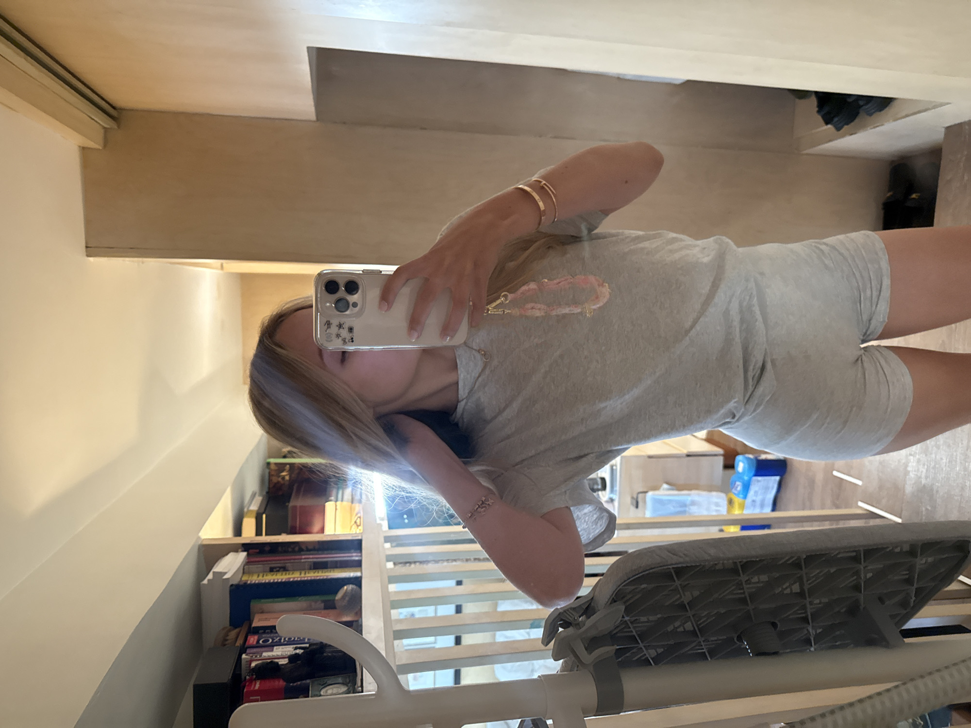 A young Caucasian woman with blonde hair, wearing a light grey t-shirt and matching grey shorts, stands in front of a mirror, taking a selfie with her smartphone. The phone has a light-colored case with a pink and white beaded charm attached to its charging port. She is also wearing two gold bracelets on her right wrist. The background shows a bookshelf filled with books on the left, and a wooden-paneled wall on the right. In the reflection, an ironing board can be seen on the left, and a bathroom or closet area with shelves and laundry detergent bottles in the background. The lighting is bright, suggesting it is daytime. The setting appears to be a domestic interior, possibly a bedroom or dressing area. There is no indication that this location is in Ciudad de México, Mexico.