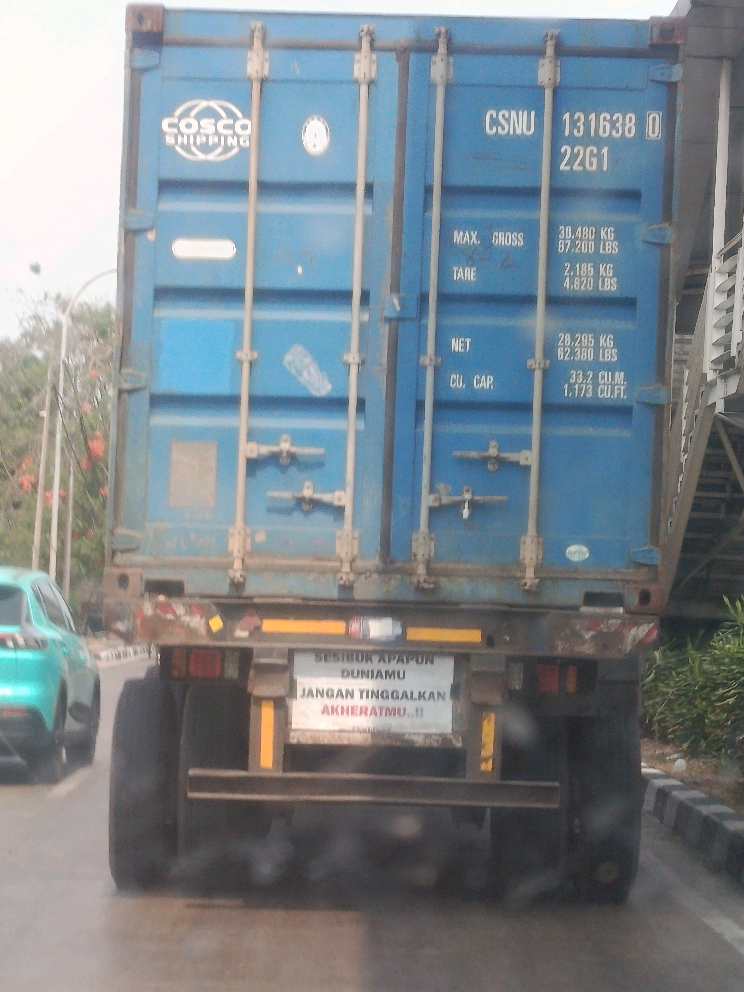 The image shows the rear of a blue shipping container on a truck. The container has markings indicating it is owned by CSNU, with the number 131638 and the code 22G1. Below this, there are weight specifications for MAX. CROSS, TARE, and NET. The container also has the COSCO SHIPPING logo on the left side. There is a sign on the back of the truck with text that appears to be in Indonesian, reading "SESIBUK APAPUN DUNIAMU JANGAN TINGGALKAN AKHERATMU..!!". A teal colored car is visible to the left of the truck, and the truck's exhaust is emitting dark smoke. The background shows trees, foliage, and a street with a curb. The scene appears to be a street in East Jakarta, Indonesia, based on the context provided in the prompt.