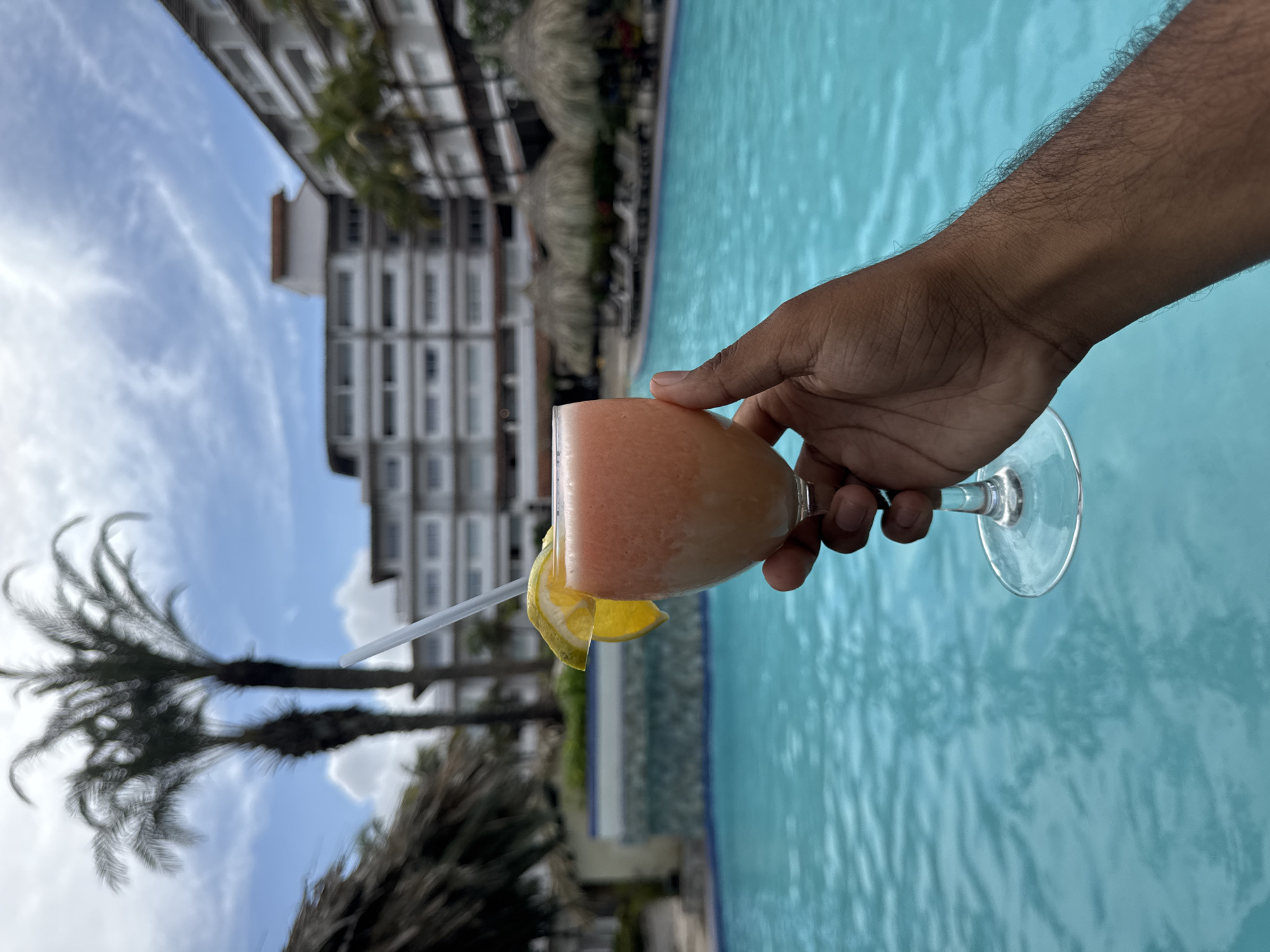 The image captures a vibrant, sunny scene, likely from a vacation in El Tirano, Venezuela. In the foreground, a person's hand, with dark skin and visible arm hair, holds a stemmed cocktail glass. The glass contains a light orange or pink frozen drink, garnished with a yellow citrus slice (lemon or orange) on the rim and a white plastic straw.
The dominant feature in the midground, spanning the right side of the frame, is a clear, inviting blue swimming pool with subtle ripples on its surface, indicating movement of water.
In the background on the left, several tall palm trees with visible fronds are silhouetted against the sky. Behind the trees, a multi-story white building with numerous windows, possibly a hotel or resort, stands under a bright blue sky with scattered white clouds. The overall impression is one of relaxation and leisure, suggesting a pleasant day spent by the pool. No visible text is present in the image.