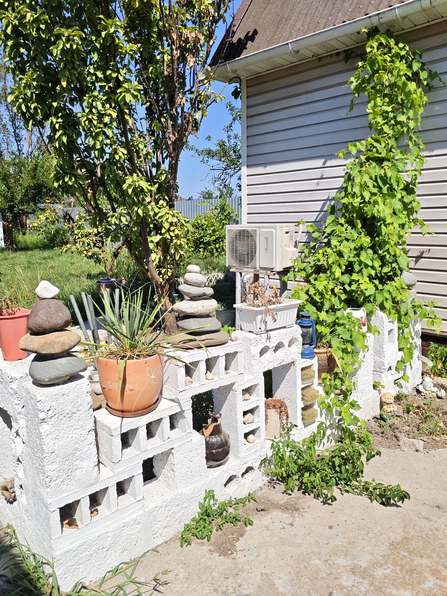 This image depicts a sunny outdoor scene in Rostov-on-Don, Russia, focusing on a decorated cinder block structure in front of a house. The structure, painted white, features stacked stones on top of it and at the base. A terracotta pot with a spiky green plant sits on the structure, and to its right, a large vine with green leaves is growing up the side of the house. An air conditioning unit is mounted on the exterior wall of the house, and a blue lantern is placed on the cinder block structure. The ground is a mix of concrete and soil, with some green plants growing. In the background, trees and a fence are visible. No people are present in the image.