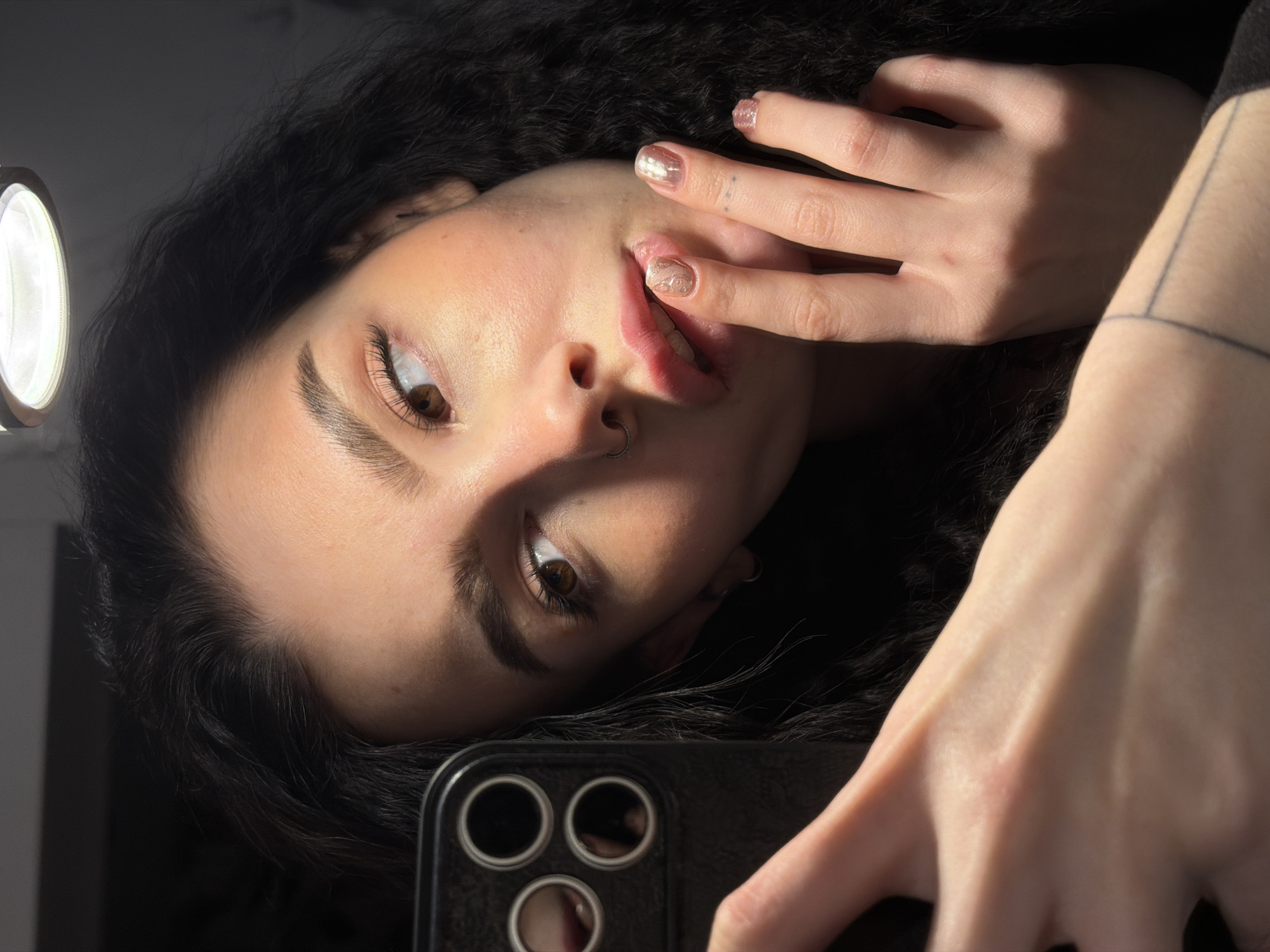 A woman with dark, curly hair is holding a smartphone and taking a selfie. Her left hand is touching her lips, and she has a nose ring in her left nostril. She has light skin and brown eyes. A ring light is visible in the upper left corner of the image. There is a tattoo on her right wrist. The photo does not appear to be set in the city of Kazan, Russia, as there are no landmarks or other features that would indicate this location.