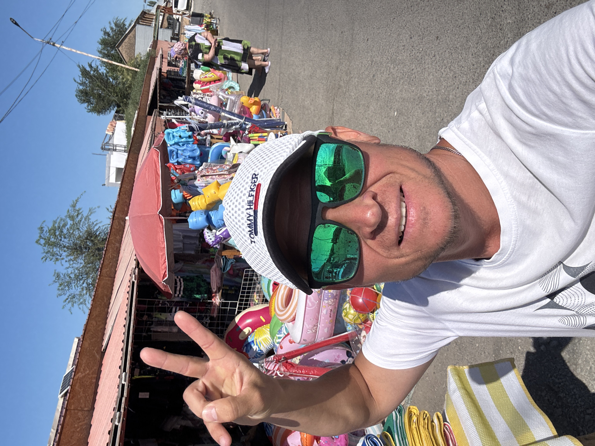 The image shows a man in the foreground taking a selfie. He is wearing a white t-shirt, a white baseball cap with "TOMMY HILFIGER" written on it, and large, mirrored sunglasses. He is making a peace sign with his left hand. Behind him, there is a street market with stalls selling various items, including inflatable pool toys, clothing, and other merchandise. A woman in a patterned dress is visible in the background, walking away from the camera. The scene appears to be outdoors on a sunny day, with buildings and trees visible in the distance. This market is located in Urdzhar, Kazakhstan.