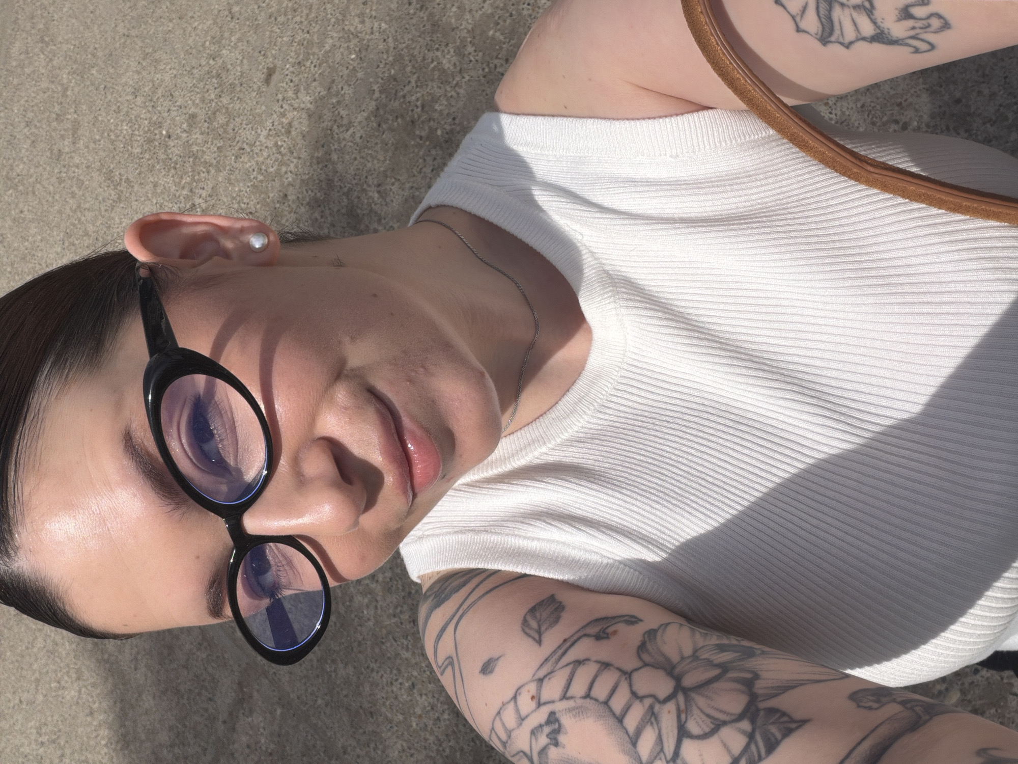 A medium, eye-level shot depicts a person with fair skin wearing black cat-eye glasses and a white ribbed shirt. The person also has tattoos on their upper arms. They are lying on a grey surface. The image does not contain explicit evidence to identify the location as Chaikovsky, Russia.