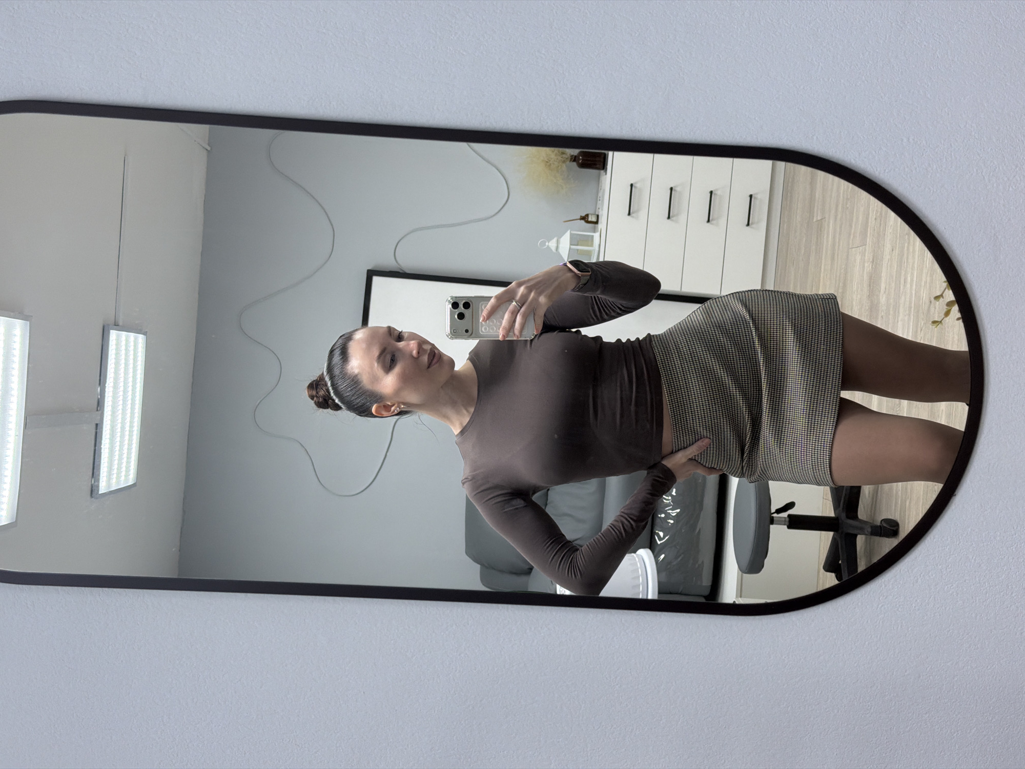 A woman in a brown long-sleeved shirt and checkered skirt takes a mirror selfie in what appears to be a residential setting. The mirror has a black frame and rounded corners. Reflected in the mirror is a room with a light gray wall, a white cabinet, and a gray chair. Two panel lights are visible on the left side of the mirror. A thin wire extends from a wall to the right of the cabinet. The woman is holding a smartphone in her right hand, and her left hand rests on her hip. The location of the city of Perm, Russia, is speculative; there are no elements within the image which can confirm the city location.