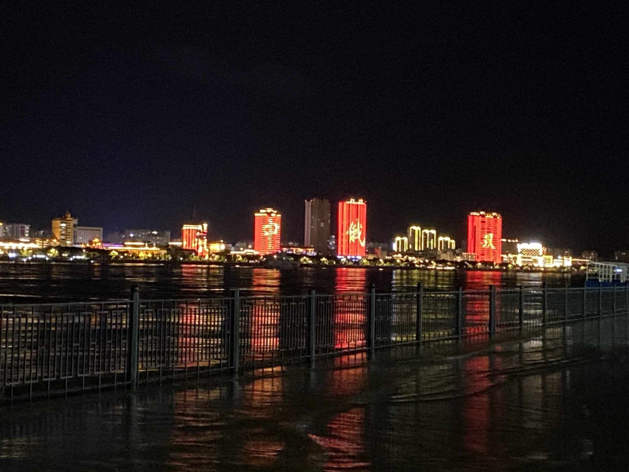 The image captures a nighttime cityscape with a body of water in the foreground, reflecting the illuminated buildings on the opposite bank. A dark metal railing runs along the edge of the water, with its reflection also visible on the wet ground. The buildings on the far side are modern high-rises, some of which are lit with bright red and yellow lights, forming patterns and characters on their facades. The sky is a deep, dark blue, suggesting it is late at night. The scene appears to be a waterfront promenade in a city.
While the image depicts a cityscape with a waterfront, it does not contain sufficient information to definitively identify the location as Blagoveshchensk, Russia. There are no visible landmarks or specific architectural styles that are uniquely associated with that city.