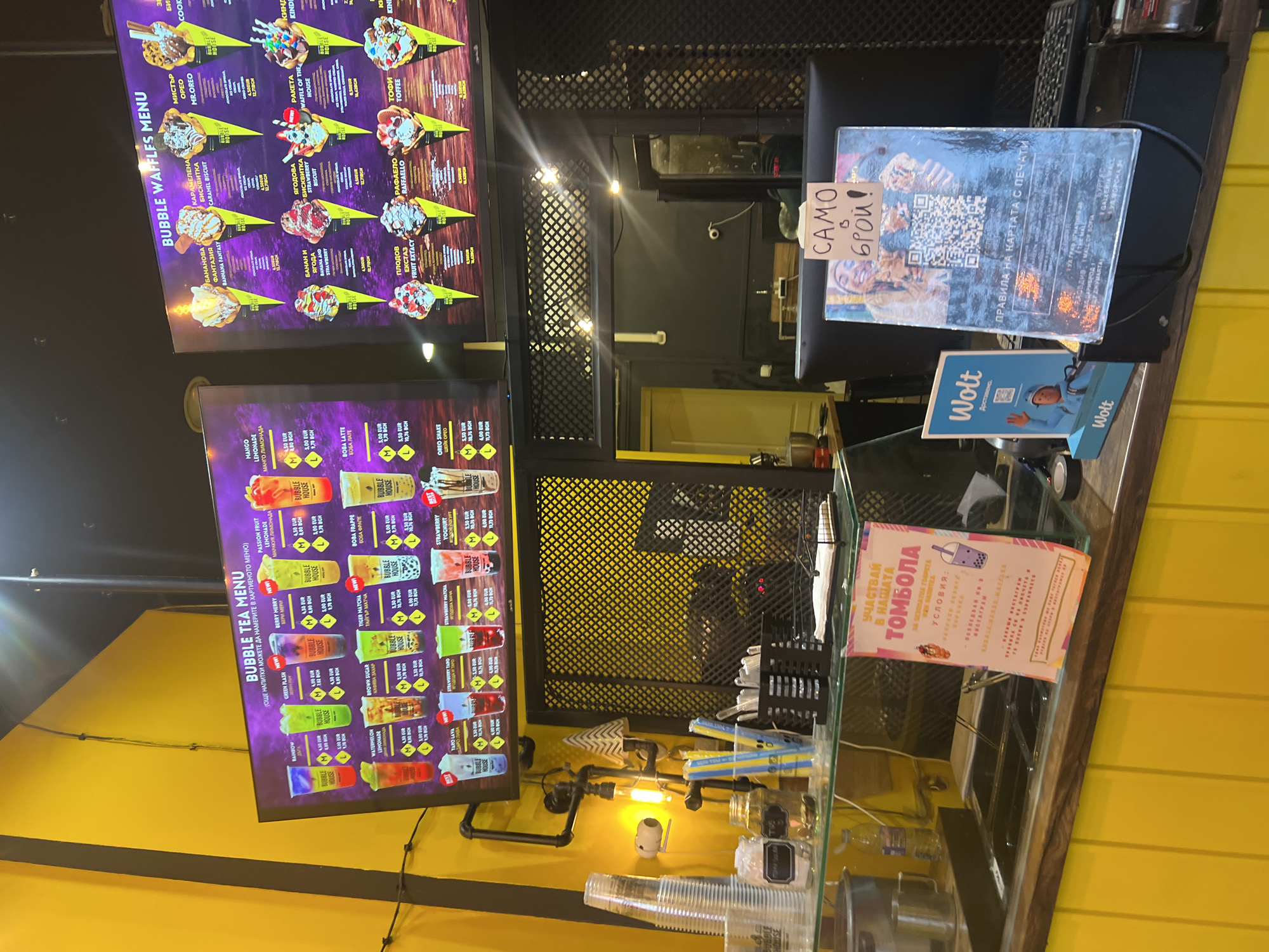 The image shows a service counter at a bubble tea and waffle shop, with menu boards displaying available items. The bubble tea menu features a variety of flavors, including passion fruit lemonade, mango lemonade, rainbow, green flash, and berry merry. The bubble waffle menu lists options such as Banana Fantasy, Caramel Biscuit, Banana and Strawberry, and Strawberry Biscuit. Promotional material includes a sign for a raffle (томбола in Bulgarian), a Wolt delivery service advertisement, and a card indicating rules, including the location: "Пловдив, Mall of Sofia" (Plovdiv, Mall of Sofia), but this is likely a mistake because Mall of Sofia is located in Sofia, not Plovdiv. There are no people visible in the image. The immediate environment includes cups, straws, and other supplies.