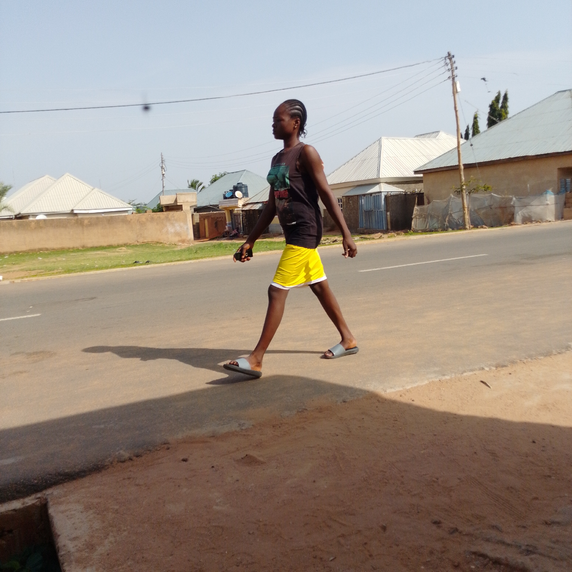 In the city of Gombe, Nigeria, a young woman with braided hair walks along a paved road. She is wearing a dark tank top and bright yellow shorts with a white hem. Her feet are adorned with gray slide sandals. She is holding a black mobile phone in her left hand. The road is lined with a low, sandy-colored wall, beyond which are buildings with light-colored metal roofs. A power pole with electrical wires is visible in the background, alongside some trees and a clear sky. The scene appears to be illuminated by daylight, casting a shadow of the woman onto the road.
