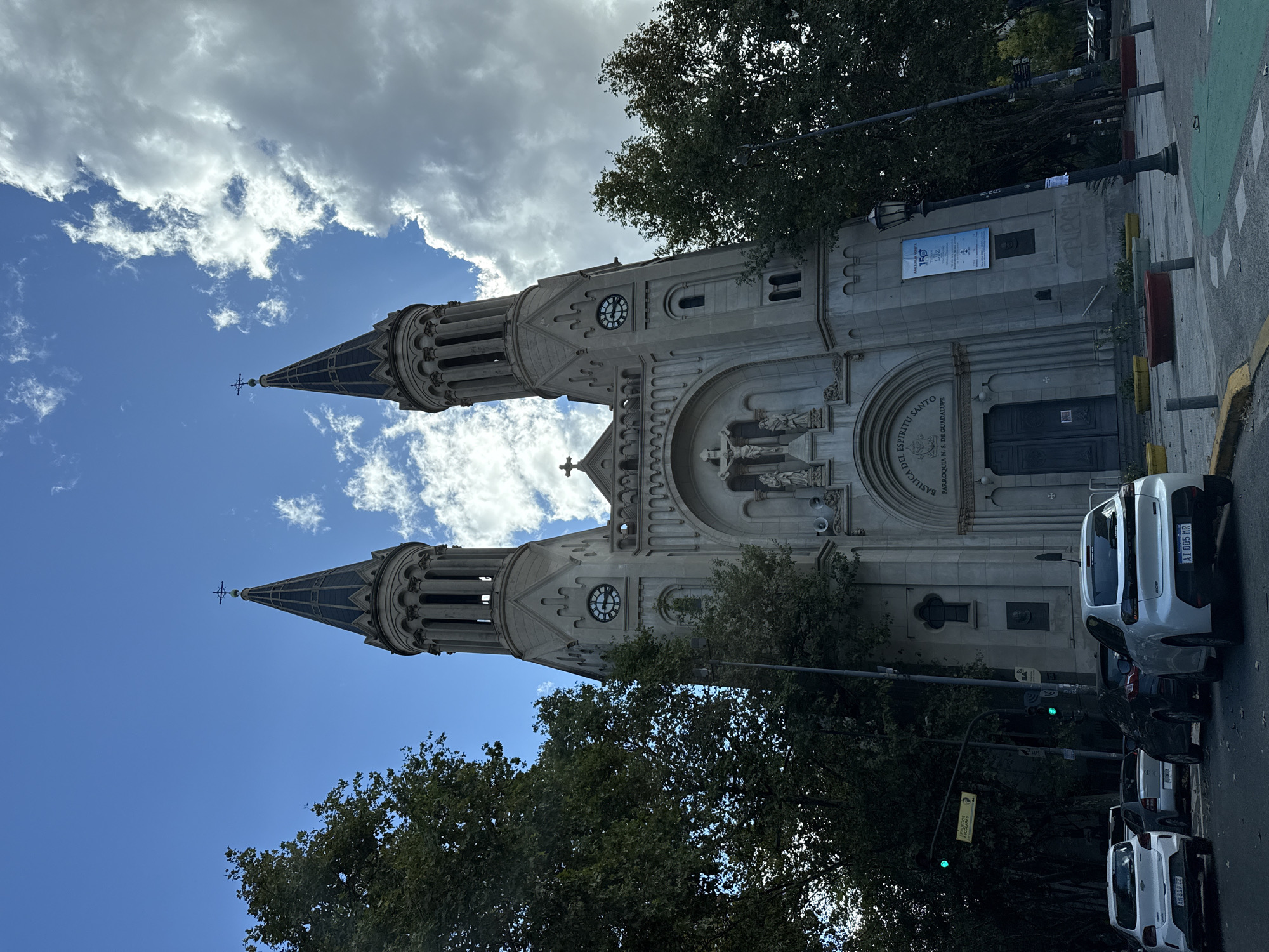 The image captures a view of the Basílica del Espíritu Santo, also known as the Parroquia Nuestra Señora de Guadalupe, in Buenos Aires, Argentina. The basilica features two tall towers with clock faces. The architecture is characterized by its Neo-Gothic style with arched doorways, statues, and decorative elements. In front of the basilica, a street with parked cars is visible. A streetlight and street signs are also present. The sky is partially cloudy, with patches of blue visible. The presence of trees partially obscures the view of the basilica. Text on the church reads BASILICA DEL ESPIRITU SANTO PARROQUIA N. S. DE GUADALUPE, and there's a posted flyer which contains the phrase "150 LÜZ". The license plate on the car nearest the church reads "AI 005 MR".