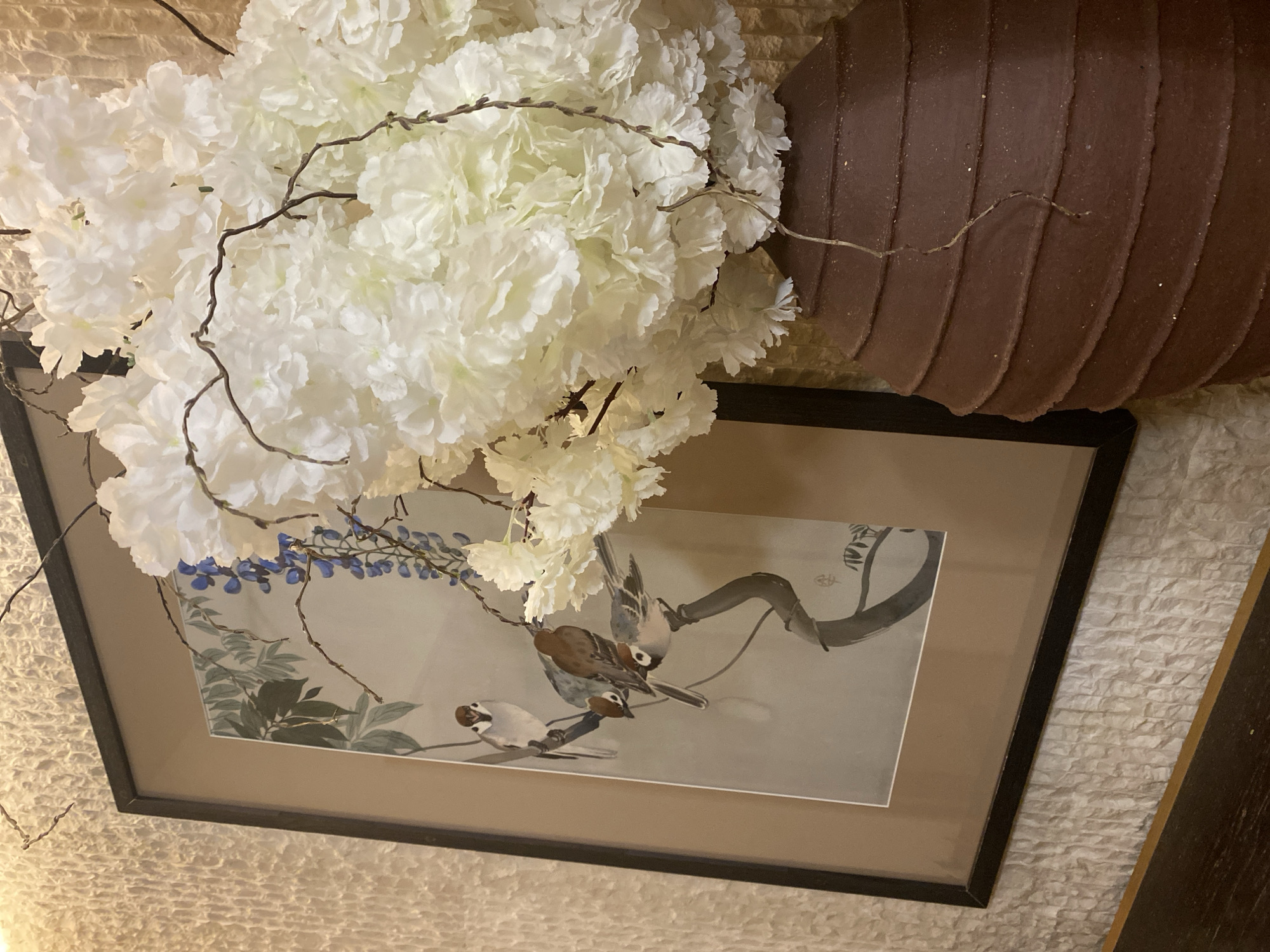 A framed artwork is mounted on a textured wall, depicting three birds perched on branches. The artwork is partially obscured by a large arrangement of white floral decorations, with some twigs extending out. To the right of the flowers is a brown, segmented cylindrical object, likely a decorative piece. The scene's location in Krasnodar, Russia cannot be determined from this single image.