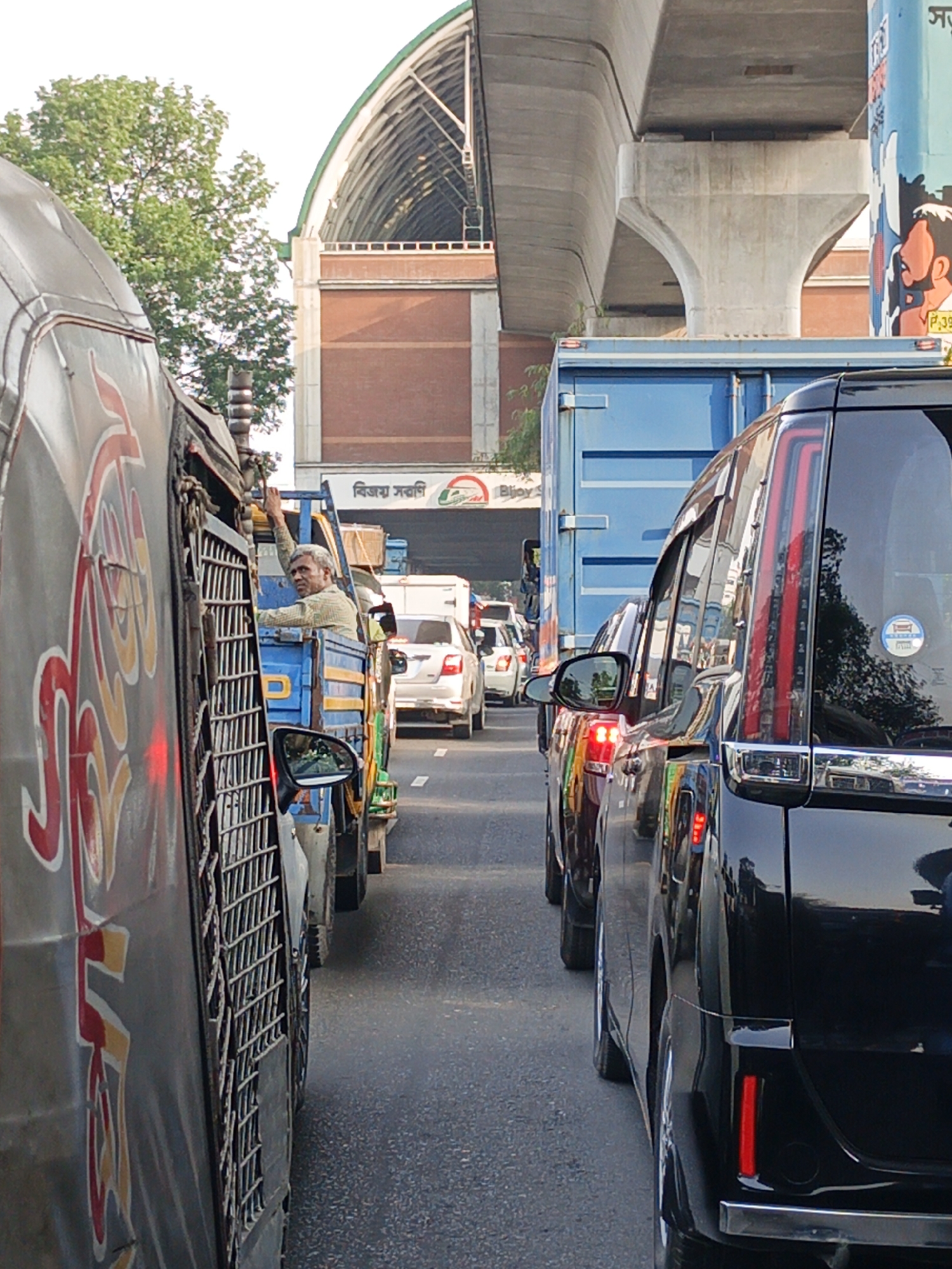 The image captures a congested street scene, likely in an urban area. Visible are several vehicles stuck in traffic, including a covered van with large red and yellow graphics, a light-blue flatbed truck with a man holding onto the cabin, a dark-colored SUV, and a blue container truck. Other cars are also visible in the distance. Above, a concrete overpass or bridge structure is present, partially obscuring the view of a building in the background. A sign is visible on the building, bearing some text in a non-Latin script. The environmental lighting suggests it is daytime. The image appears to be taken at eye-level, with the perspective of being another vehicle in the traffic.