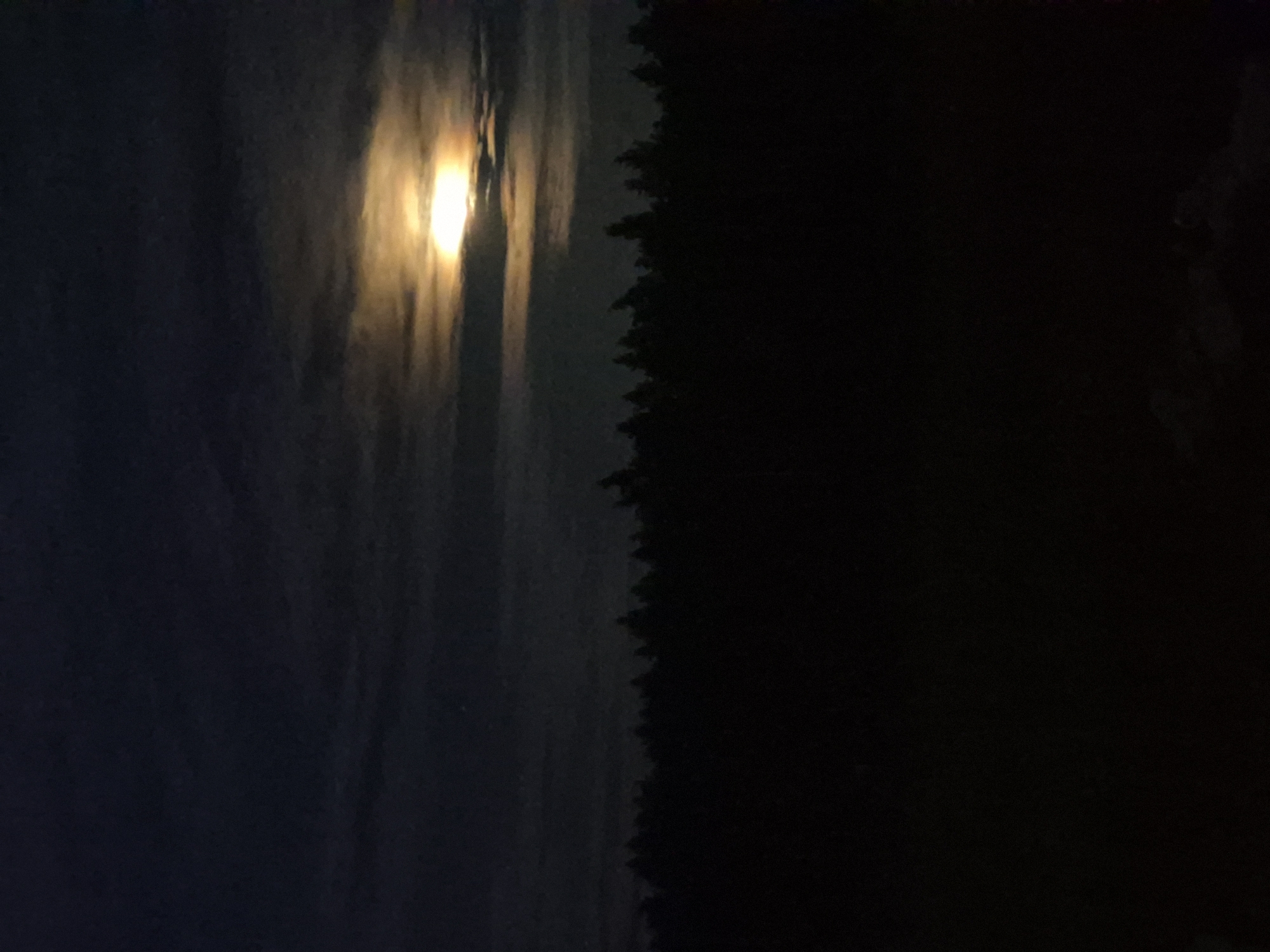 The image is a nighttime photograph of a forest edge under a cloudy sky with a bright moon. The moon is partially obscured by clouds, creating a halo effect with visible yellow and orange hues. The trees form a dark, silhouetted line against the slightly lighter sky. The foreground is dark and underexposed, with some indistinct shapes that could be ground or low vegetation. The overall impression is one of a serene, natural landscape at night. The photograph was taken in the city of Sooblase, Estonia.