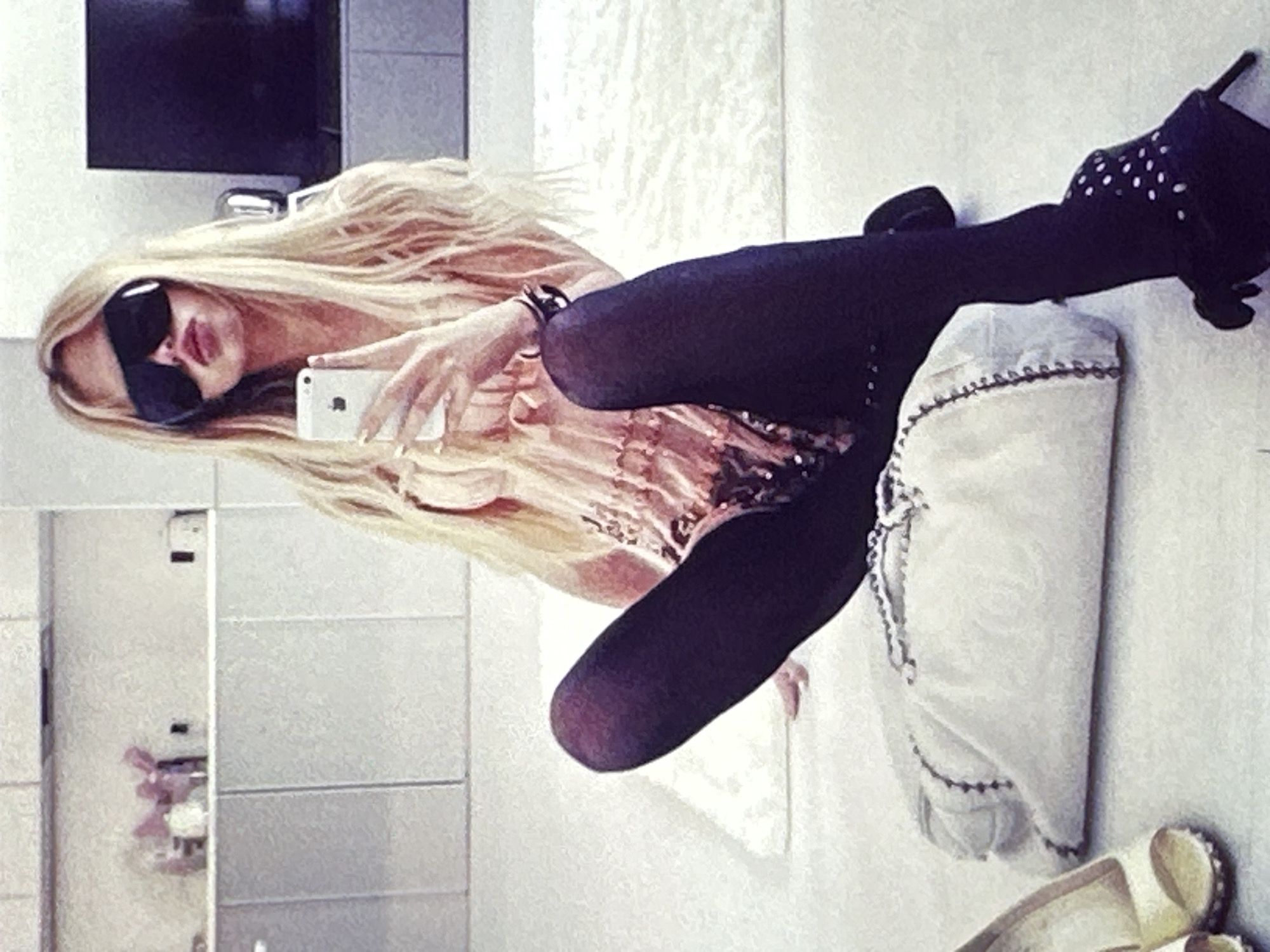 A blonde woman wearing sunglasses, a pink top, black tights, and black high-heeled boots with white dots is taking a selfie with a white iPhone. She is sitting on a white cushioned surface with a white bag beside her. Her legs are crossed, and she's holding the phone in her left hand. The background appears to be a bathroom with tiled walls and a TV screen. The location is Unknown, Unknown.