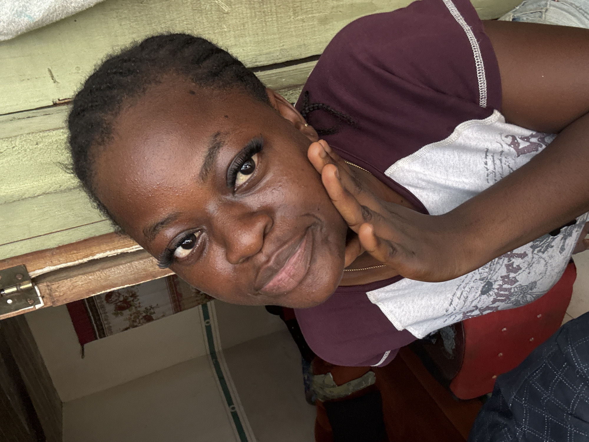 A medium close-up angled shot depicts a dark-skinned woman with false eyelashes. Her hair is dark and close to her head. She wears a maroon and white shirt and a gold necklace is visible. Her hand is touching her face. The background reveals a painted green wooden surface and interior space, possibly in a residential setting in Badagry, Nigeria. A red chair is partially visible.