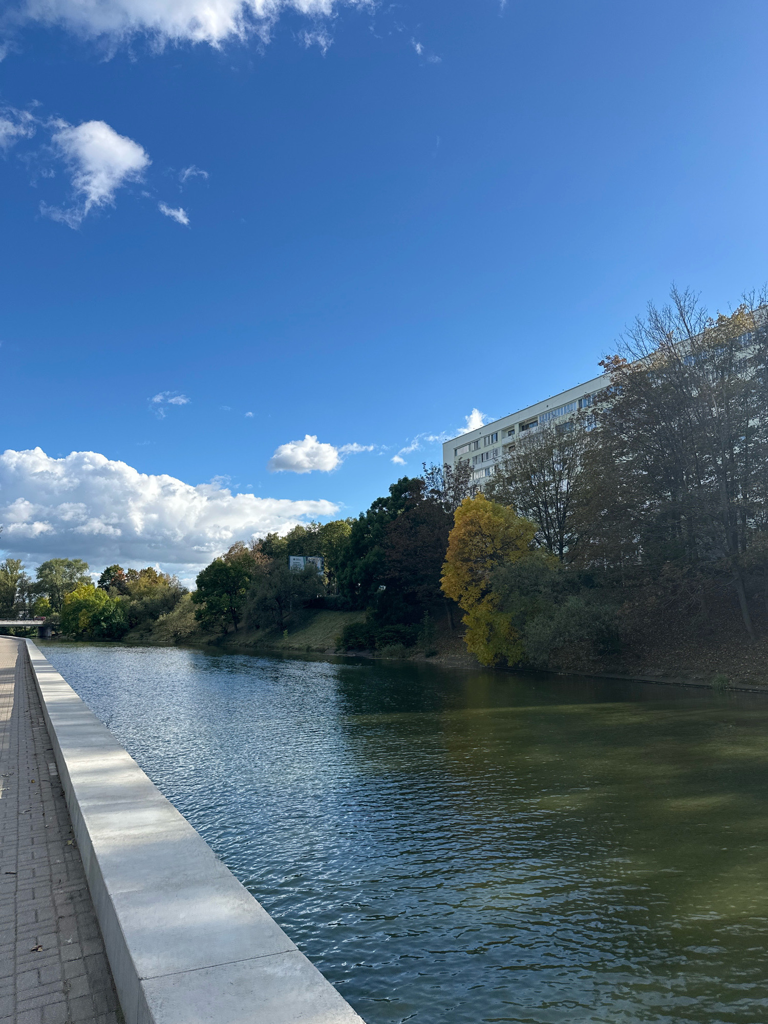 This is a photograph taken at the Svislach River embankment in Minsk, Belarus, on a sunny day with a partly cloudy sky. The image captures a wide view of the river, with a paved walkway and a stone embankment on the left. The water is calm, reflecting the blue sky and clouds. On the right side of the river, a multi-story building is visible behind a line of trees, some of which are displaying autumn foliage. The scene is serene and depicts an urban park setting.