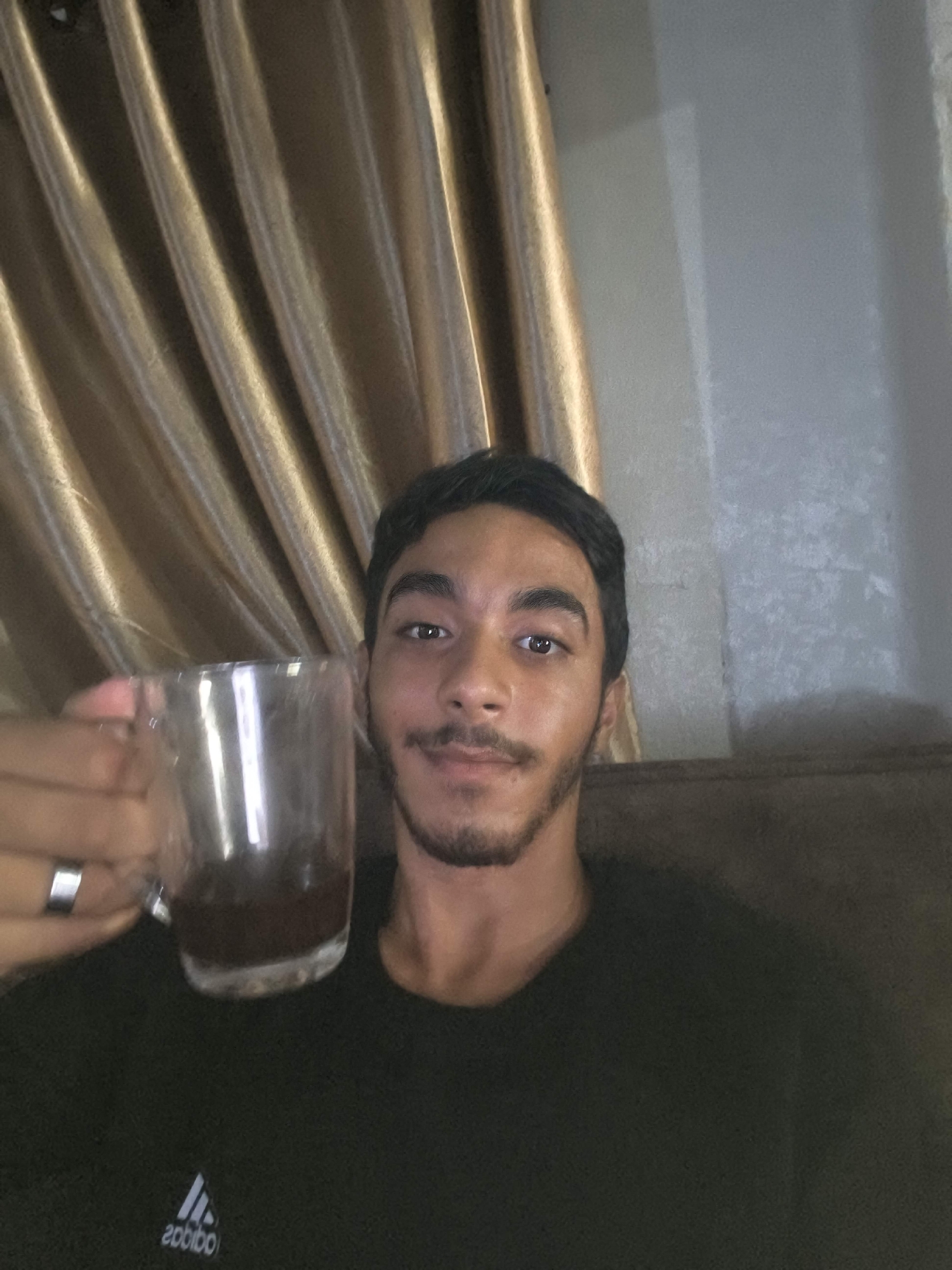 A young man with dark hair and a beard, wearing a black t-shirt with an Adidas logo, holds a glass mug of dark liquid in his left hand. He is looking at the camera and smiling slightly. The background consists of gold, pleated curtains and a textured wall. The scene appears to be indoors, possibly in a residential setting in the city of Gaza, Palestinian Territories.
