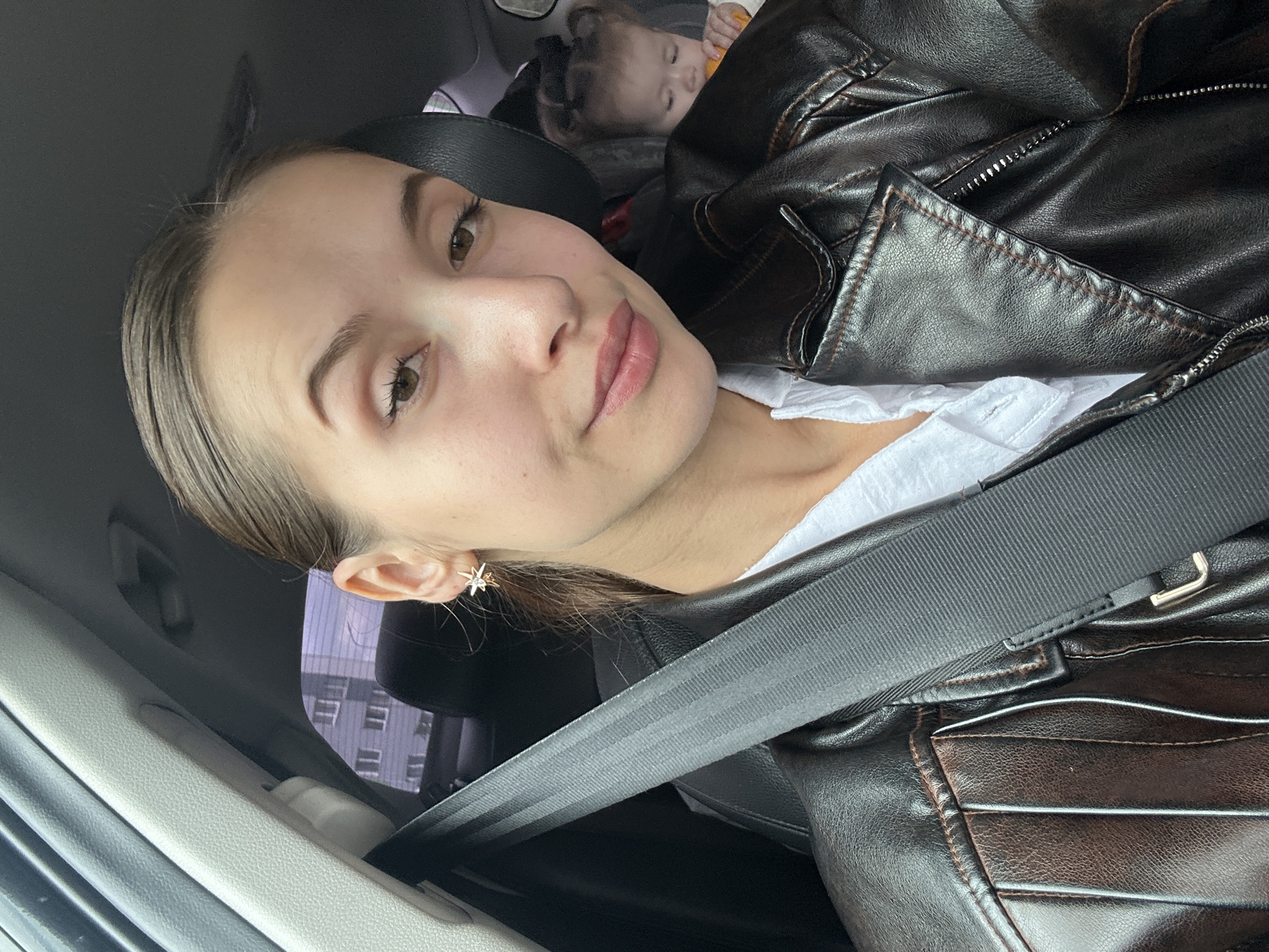 This is a selfie taken inside a vehicle. The primary subject is a young woman with her hair pulled back, wearing a dark leather jacket and a white shirt. She is looking towards the camera with a neutral expression. A seatbelt is visible across her body. In the background, a baby is visible, facing away from the camera and holding an orange object. The interior of the car is dark, with a grey headliner and a portion of a black car seat visible. Through the car window, a multi-story residential building with grey and white panels is visible, suggesting an urban setting. The overall lighting is diffused, indicating it is daytime. The presence of the building suggests the car is likely parked or moving in a residential area of Yekaterinburg, Russia, given the architectural style of the building which is common in post-Soviet cities.