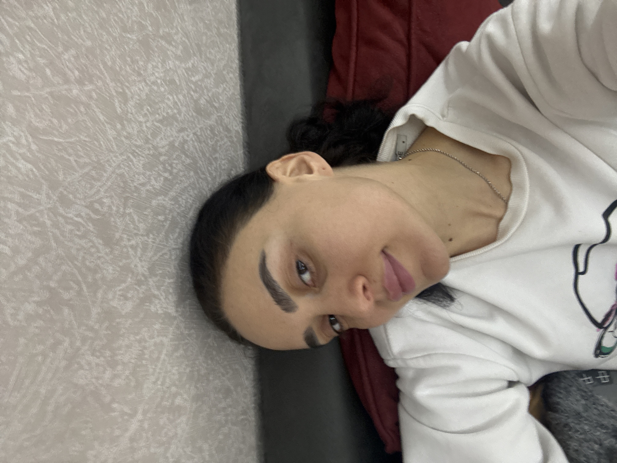 A woman is seen in a close-up shot, lying on a sofa with her head near a wall. The wall is a light grey color with a textured pattern. The sofa appears to be dark in the middle with a red cushion or backing behind her. The woman has dark hair, fair skin, and prominent eyebrows. She is wearing a white sweatshirt and a silver necklace. The specific location within Tbilisi, Georgia, is not discernible from the image.