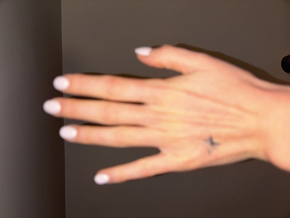 A close-up, dorsal view of a light-skinned human left hand is presented, positioned vertically with fingers slightly spread. All five fingernails are coated in a light, opaque polish, appearing white or pale pink, with an ovate shape. A small, dark, star-like or cross-like tattoo is visible on the central metacarpal area of the hand's dorsum. Subtle contours of veins and tendons are discernible on the back of the hand. The background consists of a plain, dark grey wall, with a faint horizontal seam visible in the upper portion of the frame. Illumination originates from the upper-left, casting a soft shadow to the right of the hand. A blurred, dark, unidentifiable object, potentially a person's head, is partially visible at the bottom edge of the frame. The scene is located indoors in Tunbridge Wells, United Kingdom.