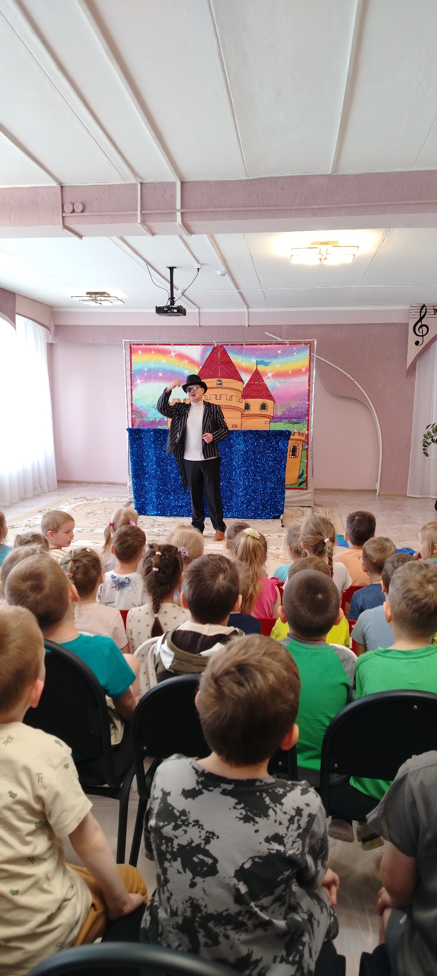 In a room in Chaikovsky, Russia, a man dressed in a black and white striped jacket, black hat, and sunglasses stands on a stage in front of a group of children. He is gesturing with his hand towards his hat. The stage has a backdrop depicting a rainbow and a cartoon castle. The children are seated in chairs and facing the stage. A projector is mounted on the ceiling above the stage. There is a white curtain on the left side of the room, and a musical note decoration on the wall on the right side of the room.