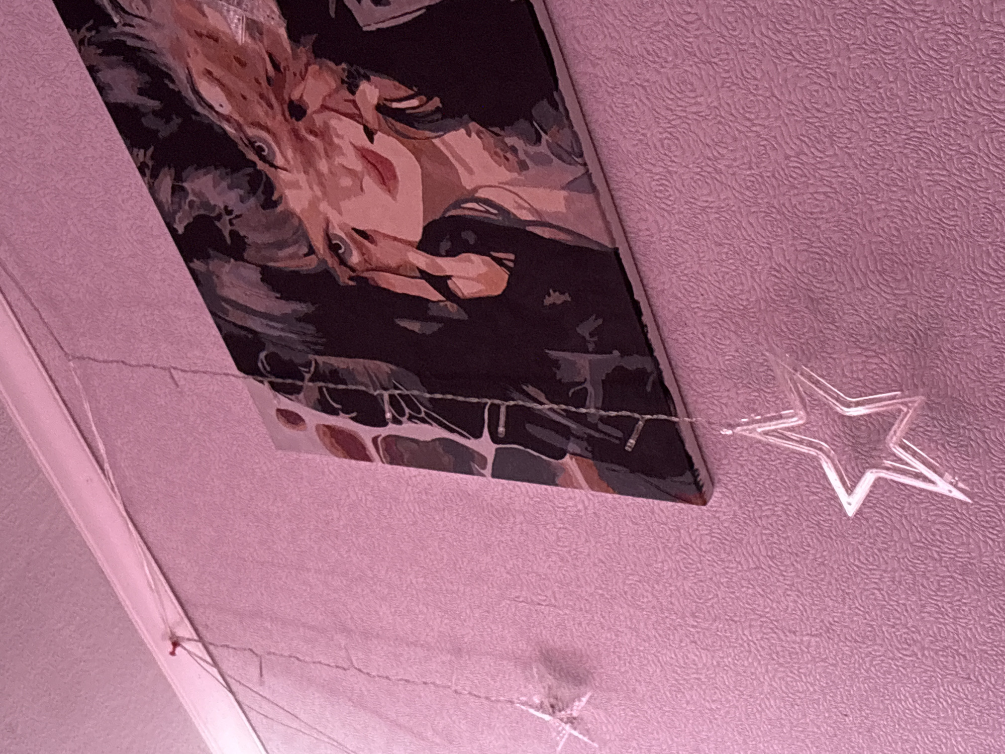 The image shows a portion of a room in Прикамский, Russia. The ceiling and part of a wall are visible, both covered in a textured pink material, possibly wallpaper. A rectangular canvas featuring an abstract painting of a woman is mounted on the wall at an angle. Below the canvas and attached to the wall is a string of lights, featuring clear plastic star-shaped decorations. One star is particularly prominent in the foreground on the right side of the frame. A white moulding is visible on the left side of the frame.