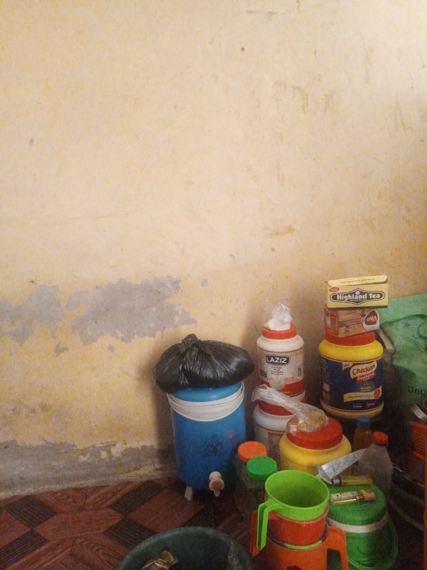 The image depicts a cluttered corner of a room with a textured, pale yellow wall showing areas of discoloration and peeling paint. In the foreground, a blue plastic water dispenser with a black plastic bag on top is positioned on a patterned floor made of brown and black tiles. To the right of the water dispenser, a stack of containers and boxes are arranged. These include a "LAZIZ" instant milk custard container, a yellow container with a red lid, a blue container labeled "Checkers Custard," and a yellow box with "Highland Tea" printed on it. Several smaller bottles and containers are also visible. Green and orange plastic cups are stacked on top of each other, with a green plastic container to their right. A green bag is partially visible in the background. The scene appears to be an indoor storage area, possibly in a home or small shop, located in Jalingo, Nigeria. There are no people visible in the image.
