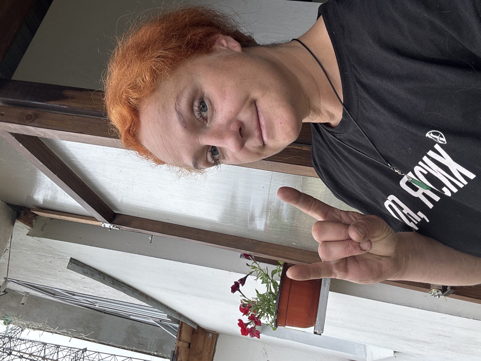 A medium shot captures a woman with bright red, curly hair posing for a selfie outdoors in Chaikovsky, Russia. She is wearing a black t-shirt and a necklace with a pendant. Her left hand is raised, forming a "rock on" hand gesture. Behind her, a white wall features a window with wooden framing and a pot of red flowers on a ledge. The overall environment appears to be a residential area, possibly a balcony or porch, with a slightly overcast sky.